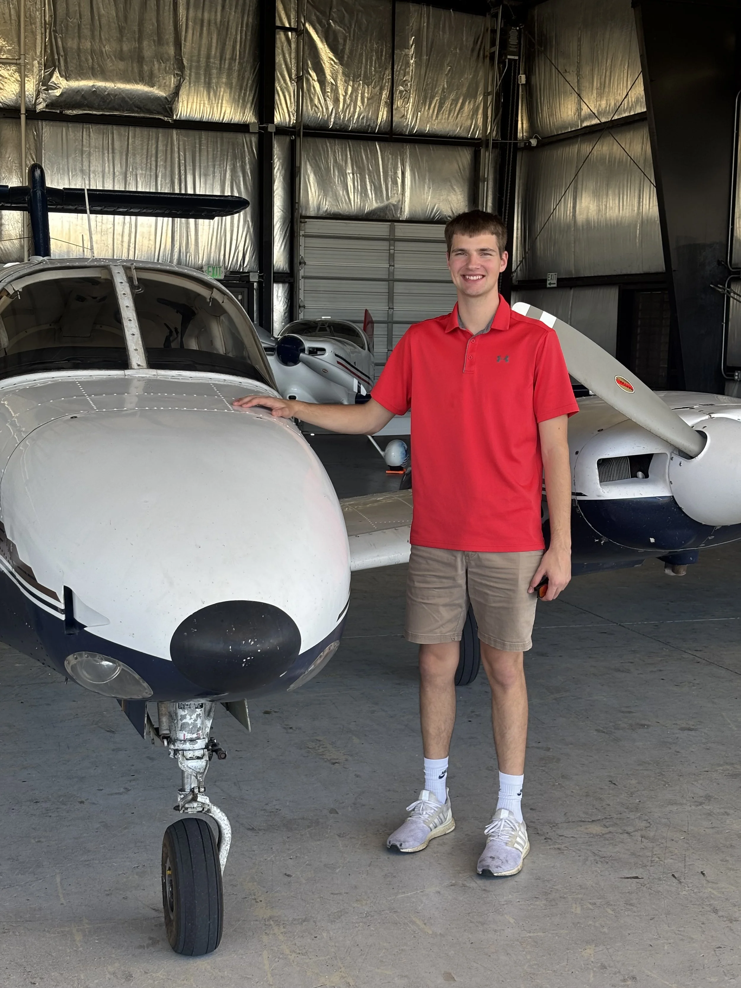 Young man in red shirt and khaki shorts standing next to a small sports plane inside an aircraft hangar.