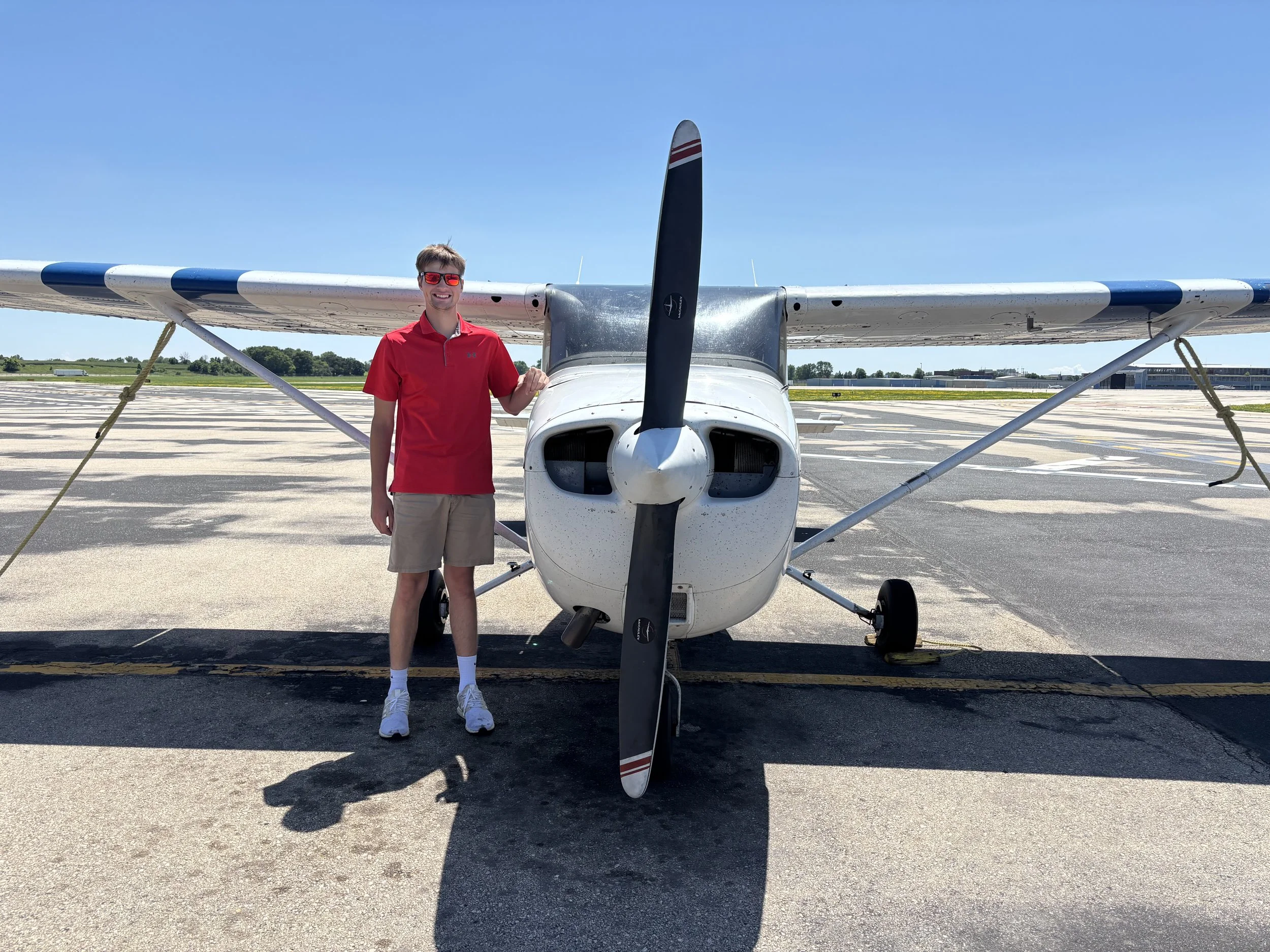 A young man in a red shirt and beige shorts standing beside a small white airplane on an airport tarmac, with a clear blue sky overhead.