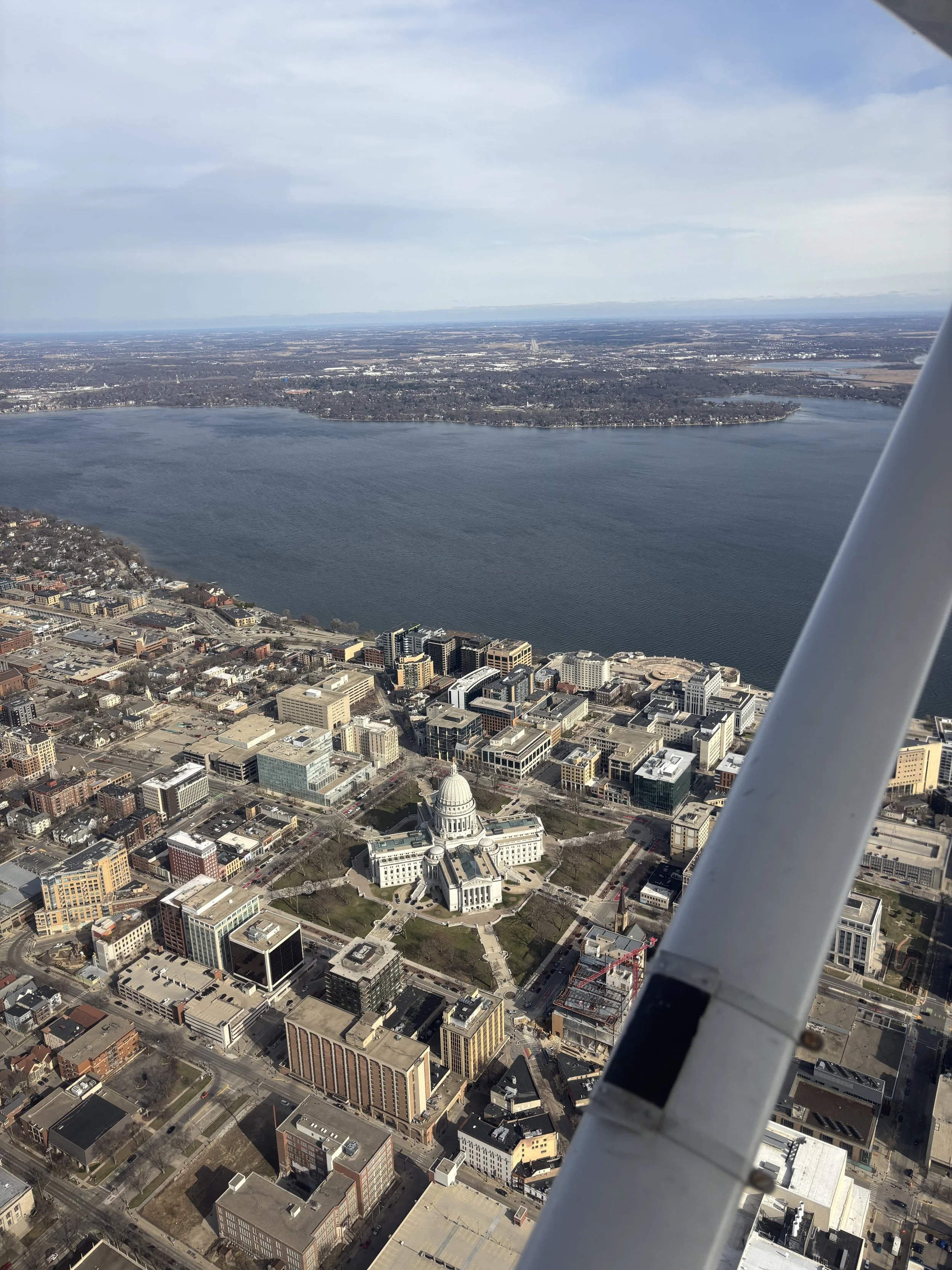 Aerial view of a city with a large river, including a prominent capitol building with a dome, and surrounding buildings under a partly cloudy sky.