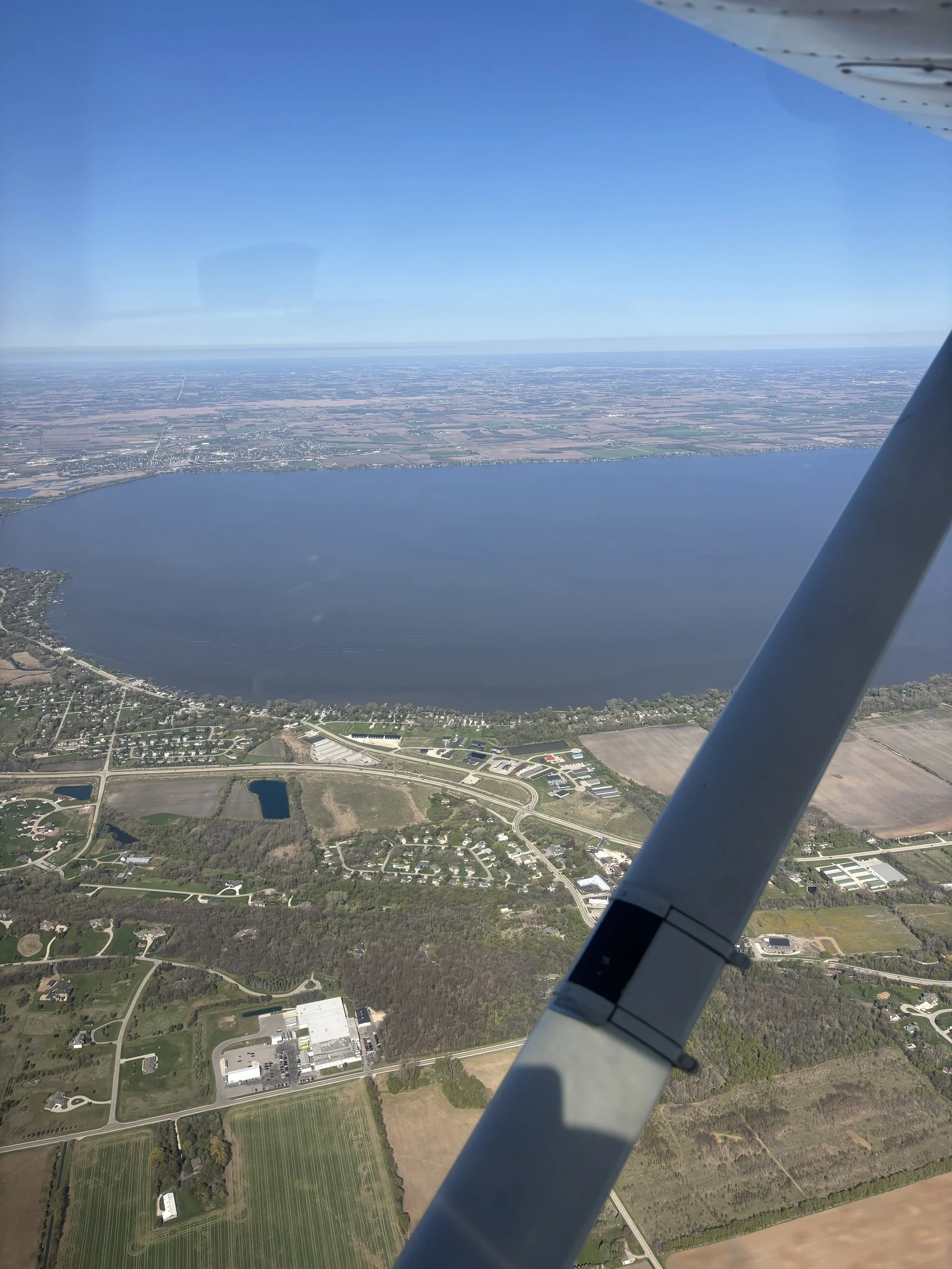 View from an airplane window shows a large lake surrounded by land with roads, buildings, and farmland underneath a clear blue sky.