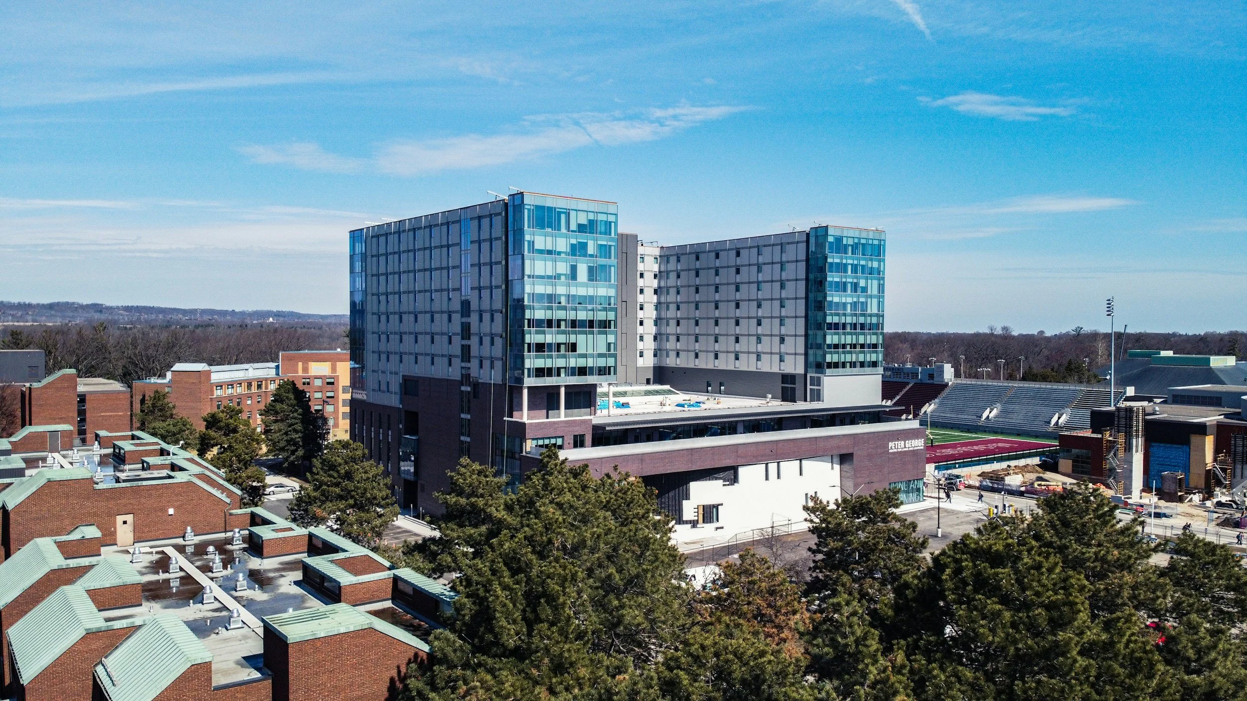 Large modern glass building on a college campus with a football stadium in the background under a blue sky.
