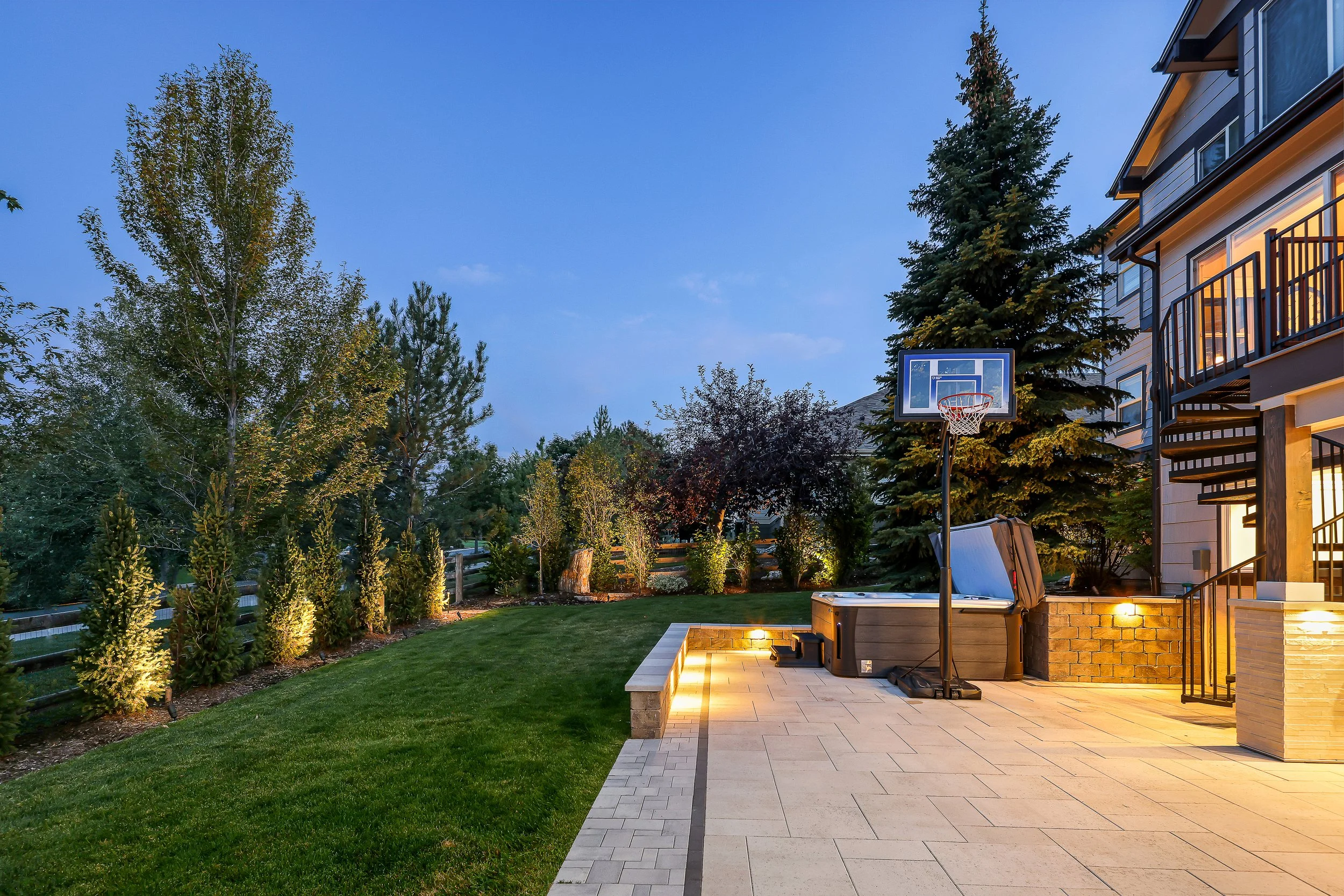 Residential backyard patio with outdoor lighting, a basketball hoop, lush green lawn, trees, bushes, and a two-story house with balconies at dusk.