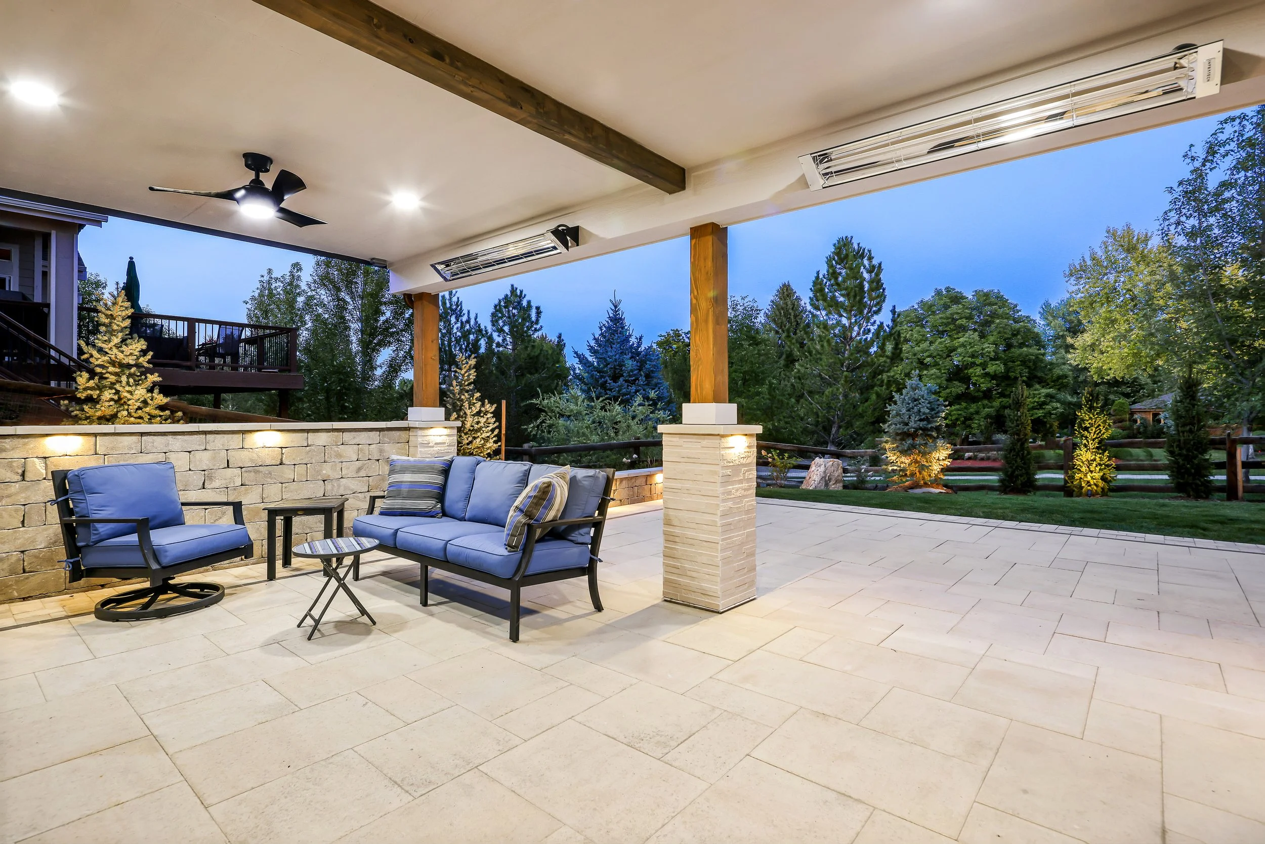 A spacious outdoor patio with seating chairs, tables, illuminated by ceiling lights, overlooking a garden with trees and a starry sky in the evening.