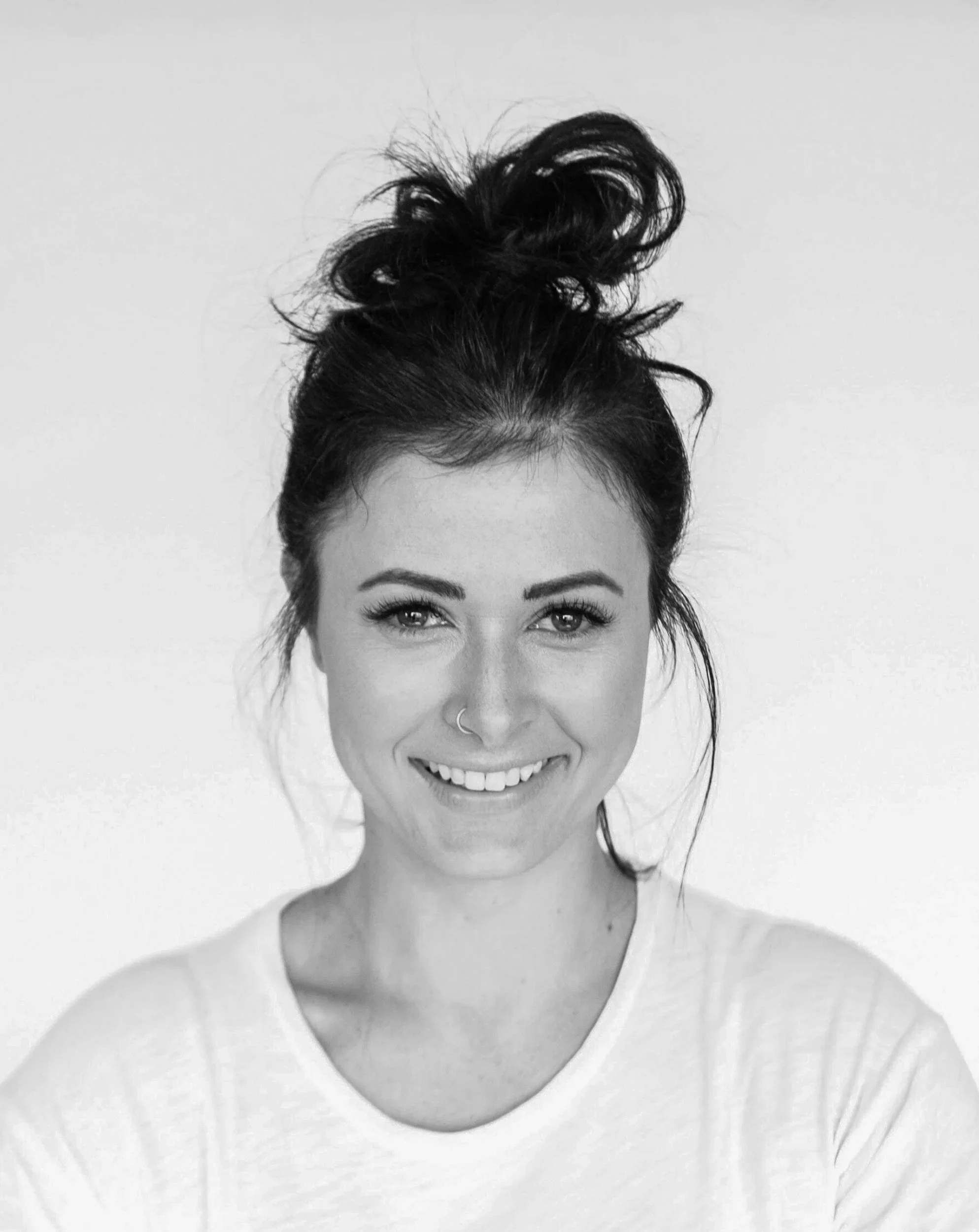 Black and white photo of a woman with a wide smile, curly hair tied up in a bun, and wearing a nose ring and a light-colored top.