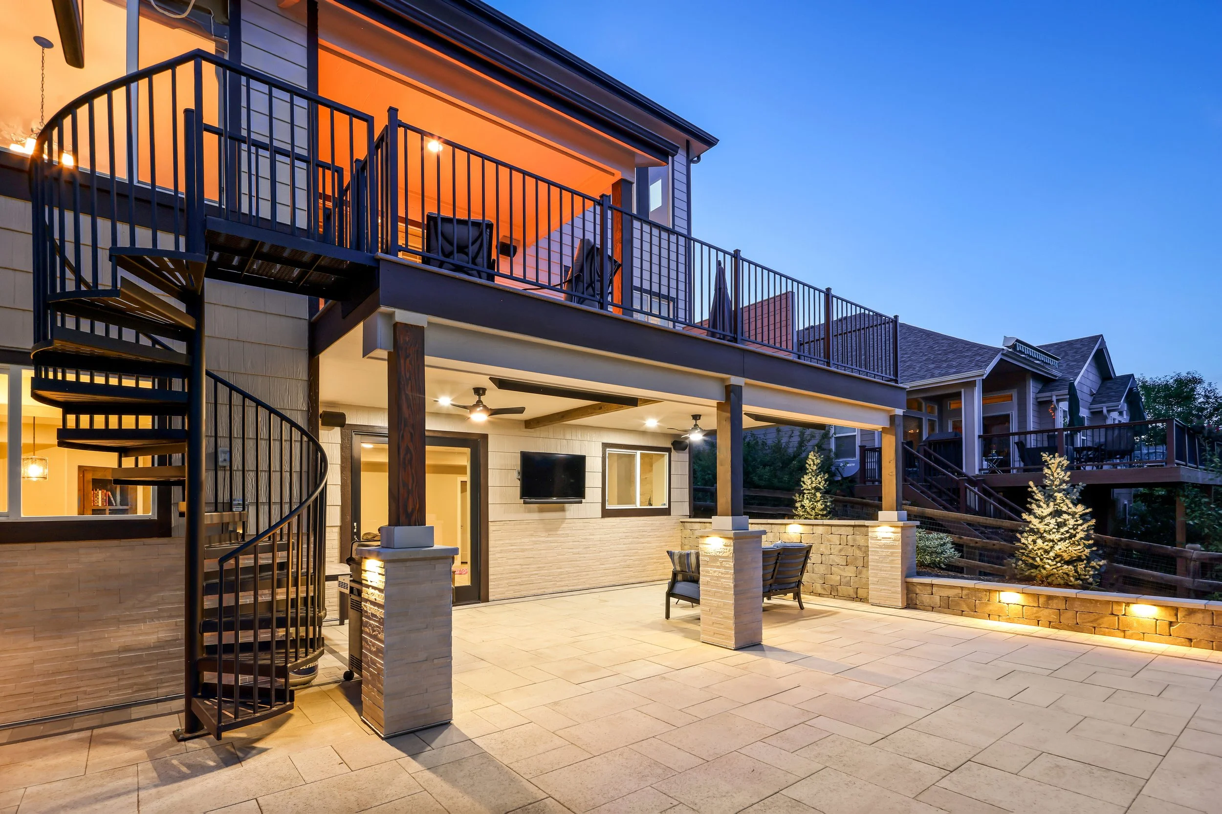 The image shows a backyard patio area of a modern house during dusk. The patio features a brick and concrete floor, outdoor furniture, a wall-mounted TV, ceiling fans, and landscaped plants. The second story has a balcony with patio furniture and umb
