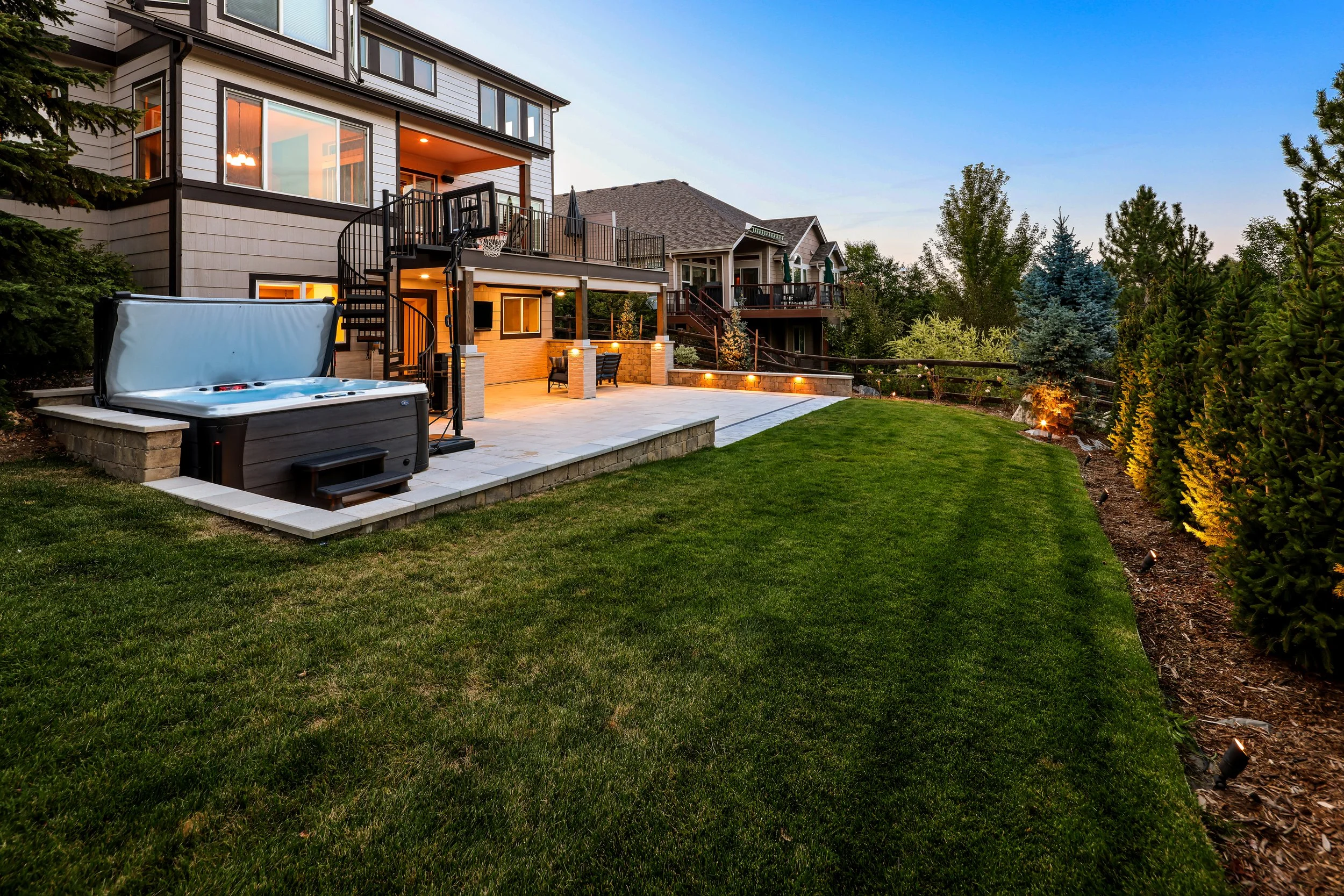 A backyard with a grassy lawn, a patio with outdoor furniture, a hot tub, and a house with multiple balconies and large windows. There are trees and shrubs around the yard, and the scene is illuminated by outdoor lighting at dusk.