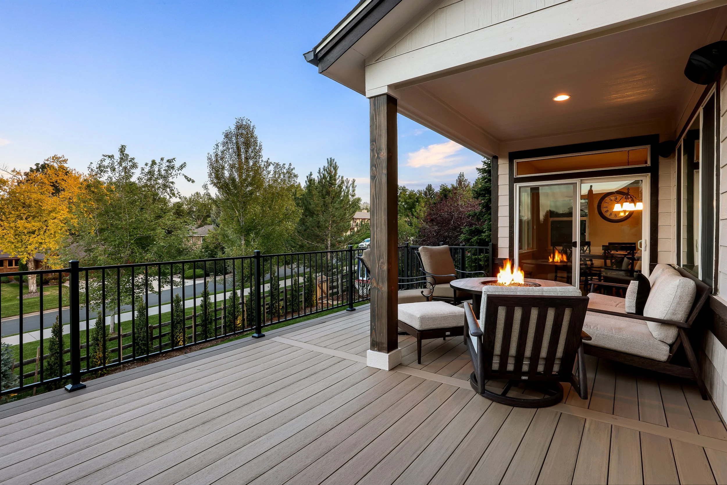 A spacious balcony with beige wooden flooring, outdoor seating including cushioned chairs and a small table with a fire pit. The balcony has black metal railing and overlooks a green landscape with trees and neighboring houses. Inside, a clock and a 