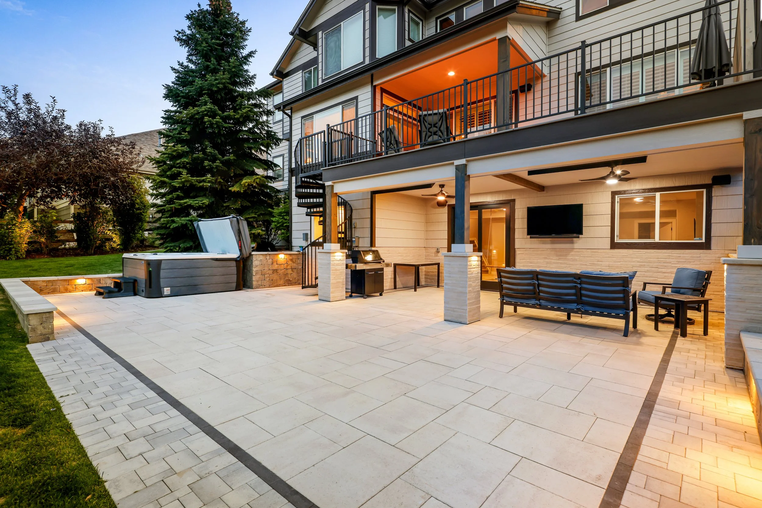 A backyard patio with outdoor furniture including a black loveseat and matching chairs, a hot tub, a built-in barbecue grill, and flat-screen TV, surrounded by a house with multiple levels, wooden siding, and large windows, with trees and lawn visibl