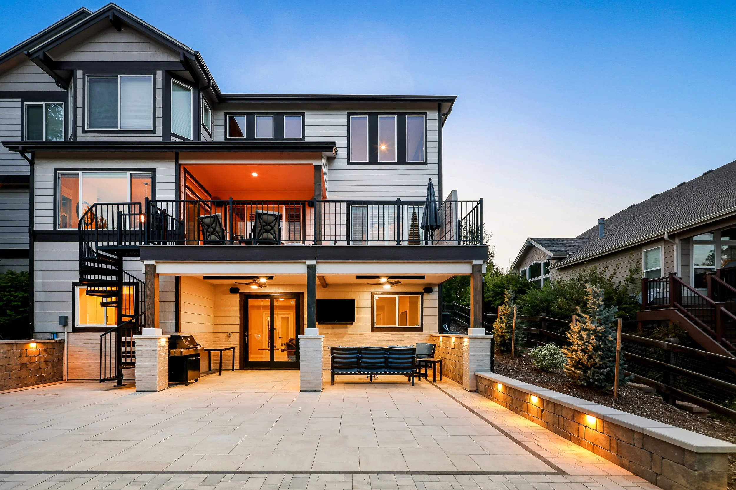 A modern multi-story house with large windows and a spacious outdoor patio area, illuminated with warm lighting at dusk.