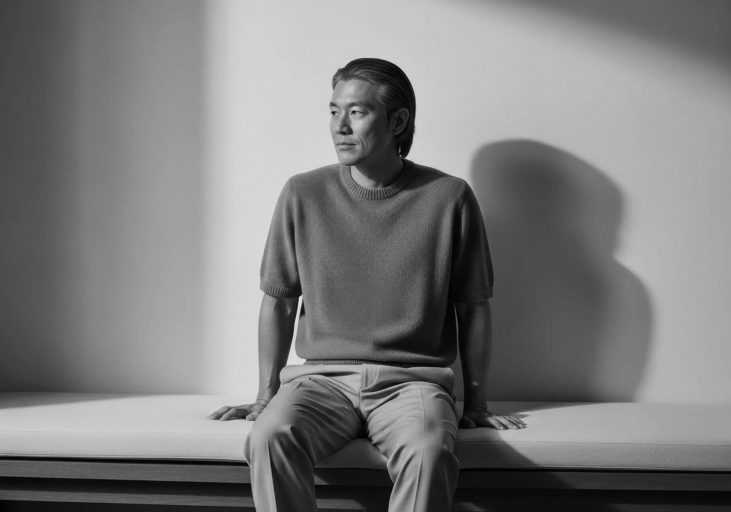 A black and white photo of a man wearing a sweater and light-colored pants, sitting on a bed with his legs crossed. He is looking to his left with a calm expression, with a plain wall behind him and a shadow cast on the wall.