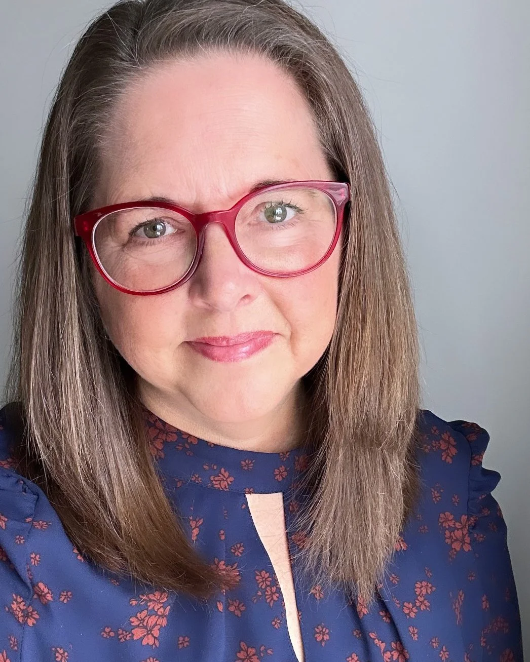 Close-up of a woman with shoulder-length brown hair, wearing pink glasses and a navy blue blouse with red floral pattern, smiling softly against a plain light gray background.