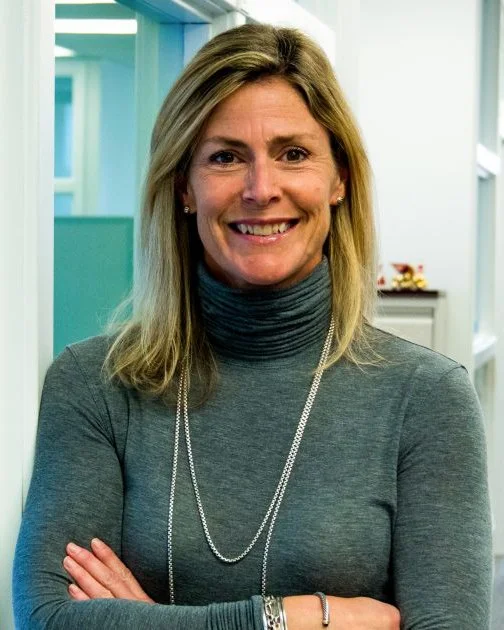 A woman with shoulder-length blonde hair wearing a gray turtleneck sweater and double-layered necklaces, smiling with crossed arms in an indoor office setting.