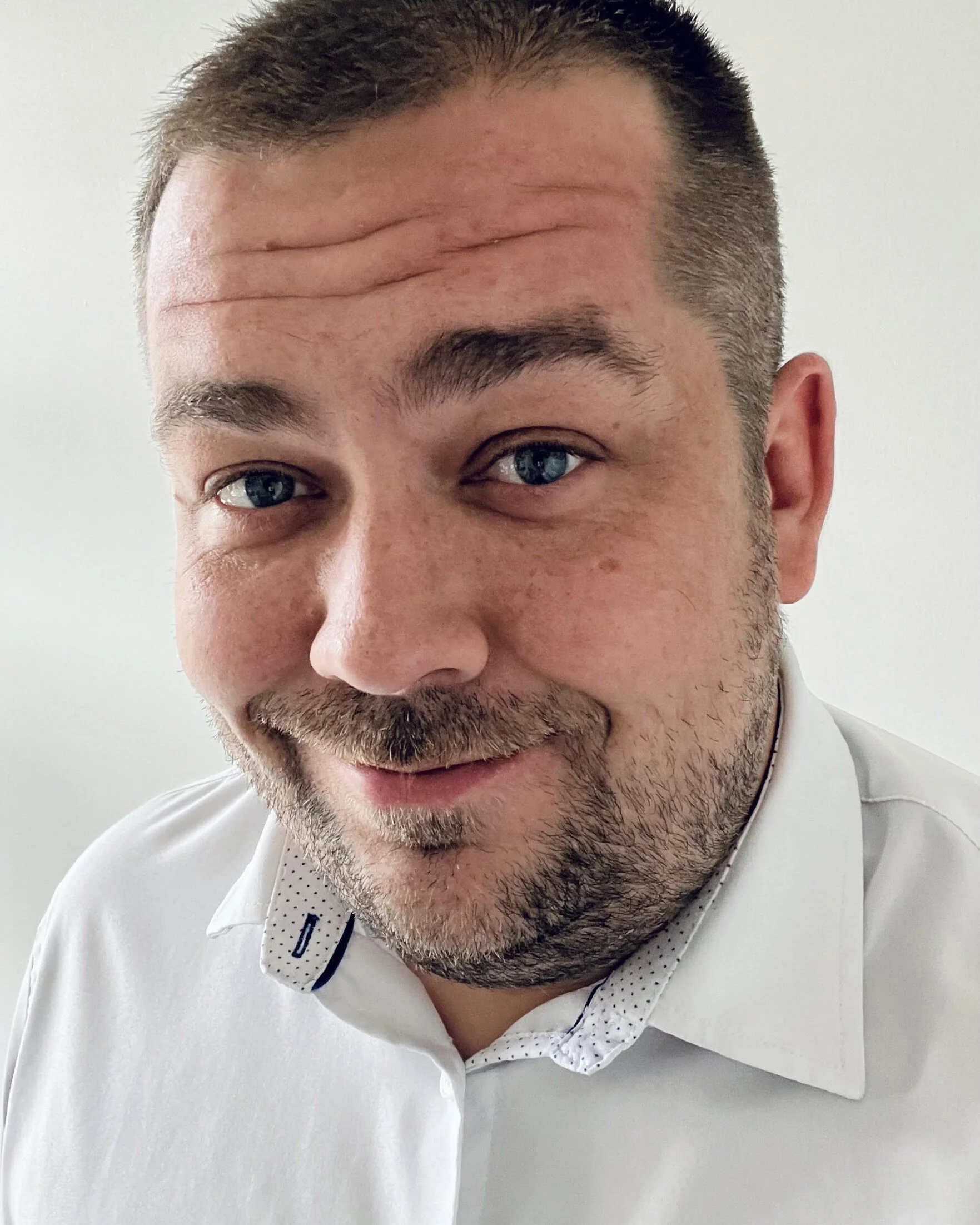 A close-up of a man's face, half smiling, with short brown hair, blue eyes, a beard, and wearing a white shirt with a dotted collar.