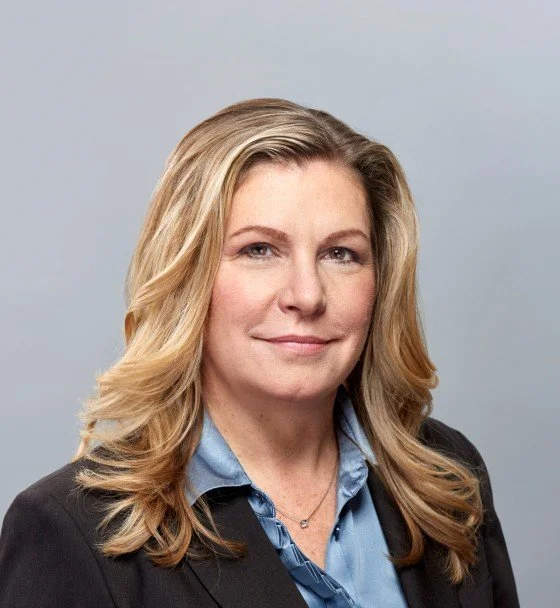 Professional woman with blonde hair wearing a black blazer and blue collared shirt, posing for a portrait against a gray background.