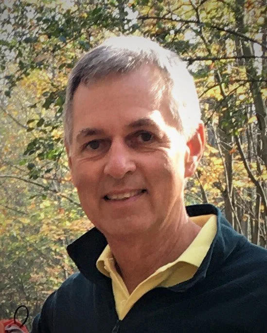 A middle-aged man with short gray hair smiling outdoors in front of trees with fall foliage.