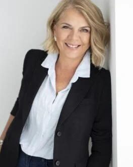 Smiling woman with blonde hair in a business suit leaning against a wall.