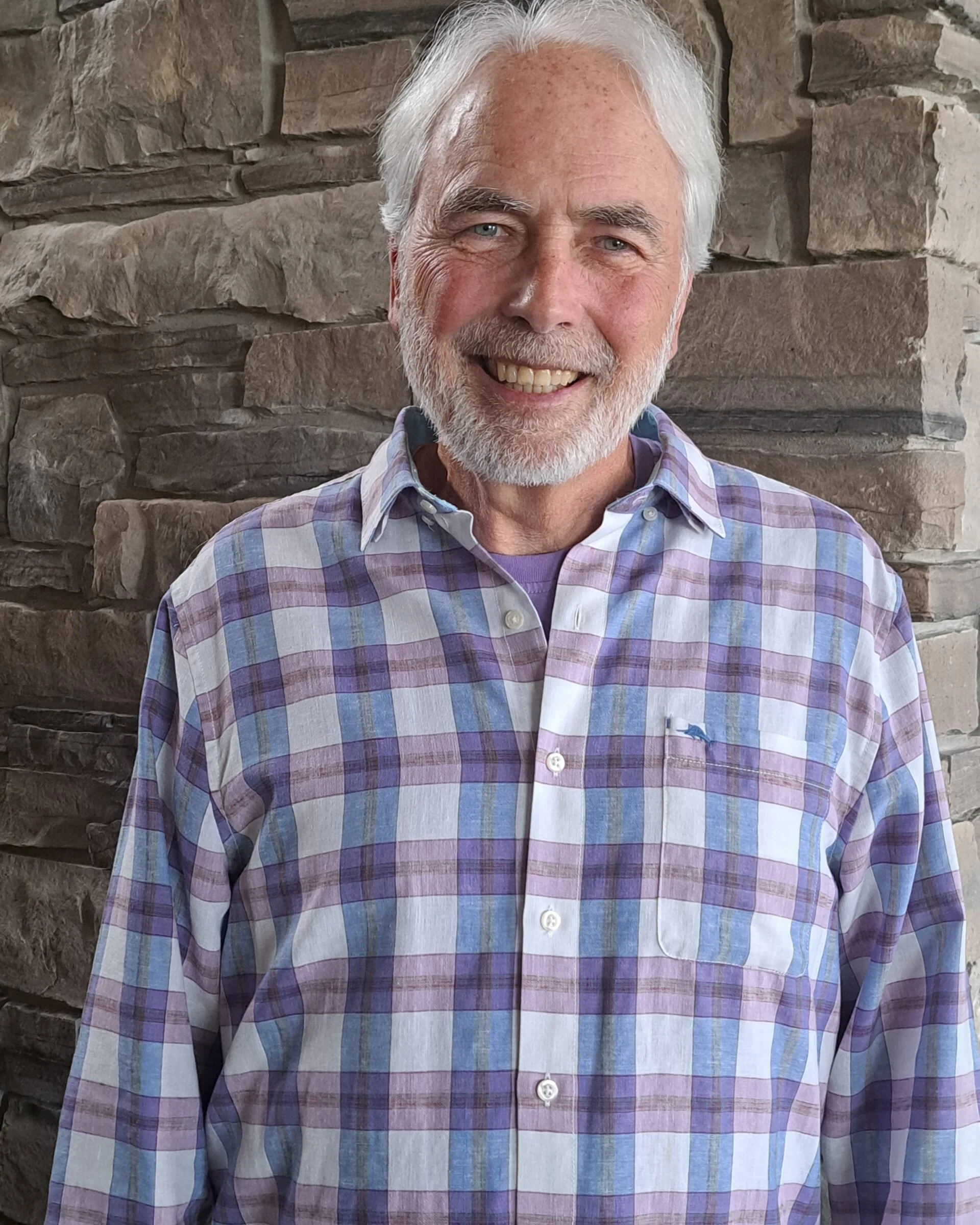 A smiling elderly man with white hair and a beard, wearing a purple and white plaid shirt, stands in front of a stone wall.