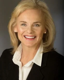 Portrait of a smiling middle-aged woman with blonde hair, wearing a black blazer and white shirt, against a neutral background.