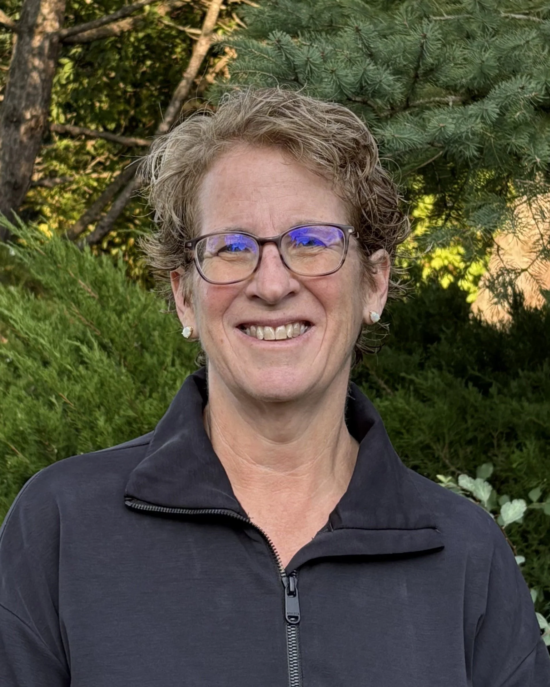 Close-up of smiling woman with short, curly, gray hair and glasses, wearing a black zip-up jacket, standing outdoors with green trees and shrubs in the background.