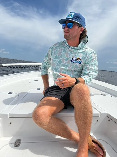 Man (pool service technician) sitting on boat deck wearing sunglasses, a blue cap, a long-sleeve shirt with a logo, and black shorts, with water and sky in the background.