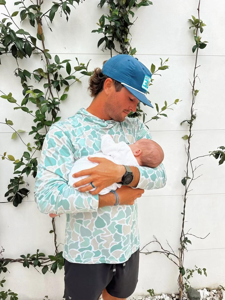 A man wearing a blue cap, patterned shirt, and a watch, holding a newborn baby wrapped in a white blanket, standing against a white wall with green vines.