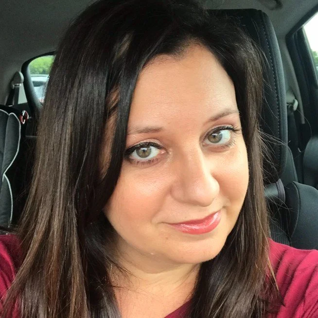 Close-up of a woman with long dark hair and light blue eyes, wearing a maroon top, sitting in a car.