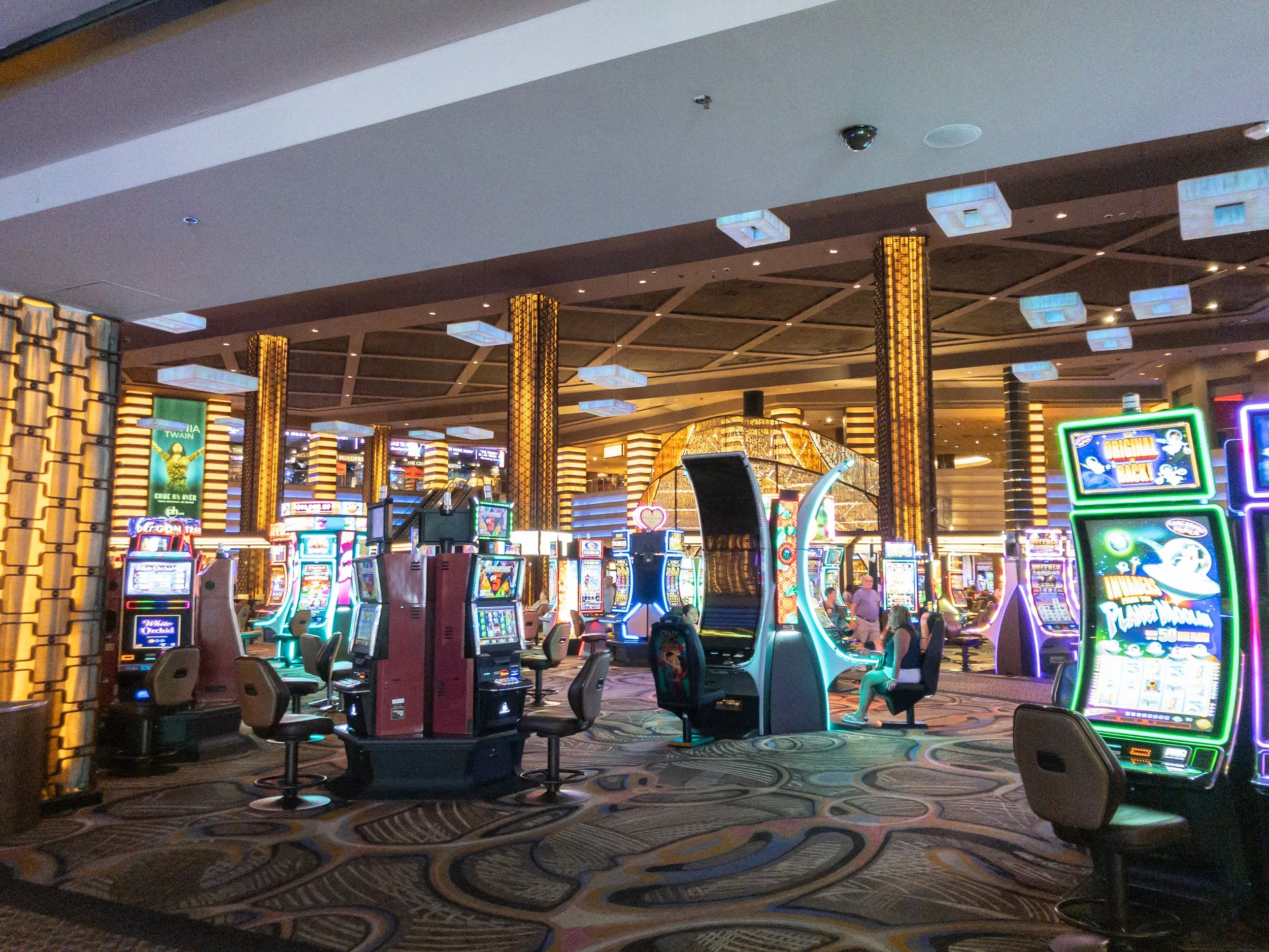 Inside a casino, slot machines and electronic gaming machines are arranged in rows, with a colorful patterned carpet on the floor. The ceiling features geometric light fixtures and decorative columns with illuminated patterns, creating a vibrant atmosphere.