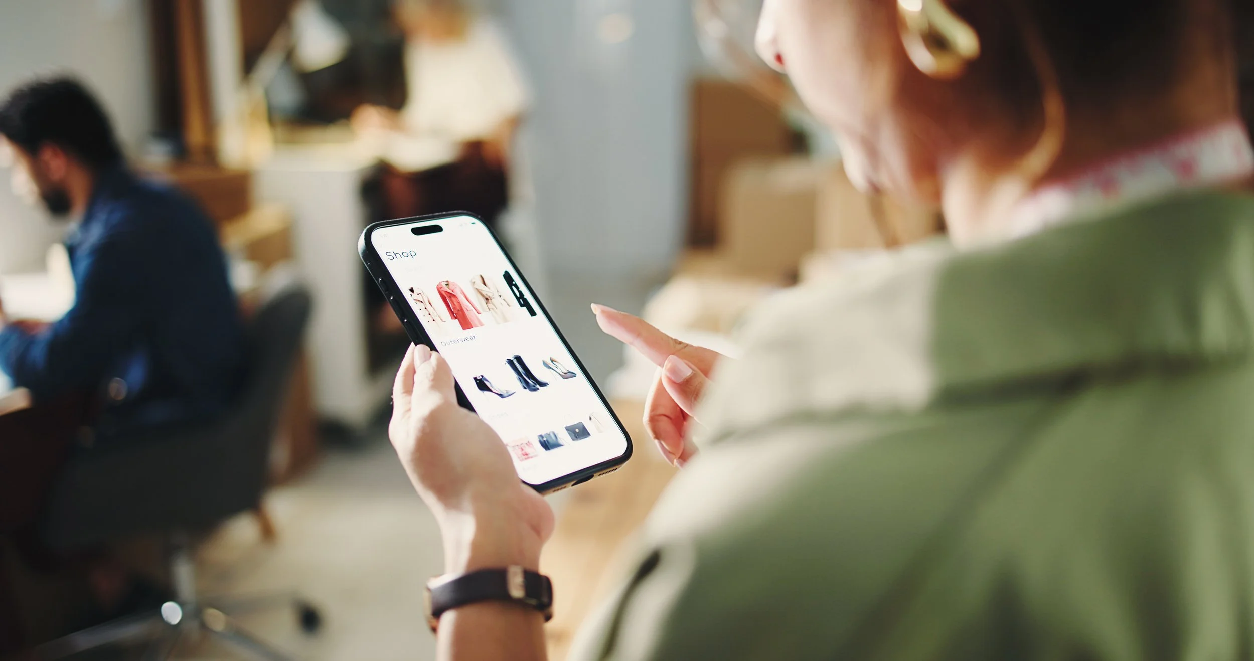 A woman holding a smartphone and shopping online for clothing items in a store, with other people in the background.