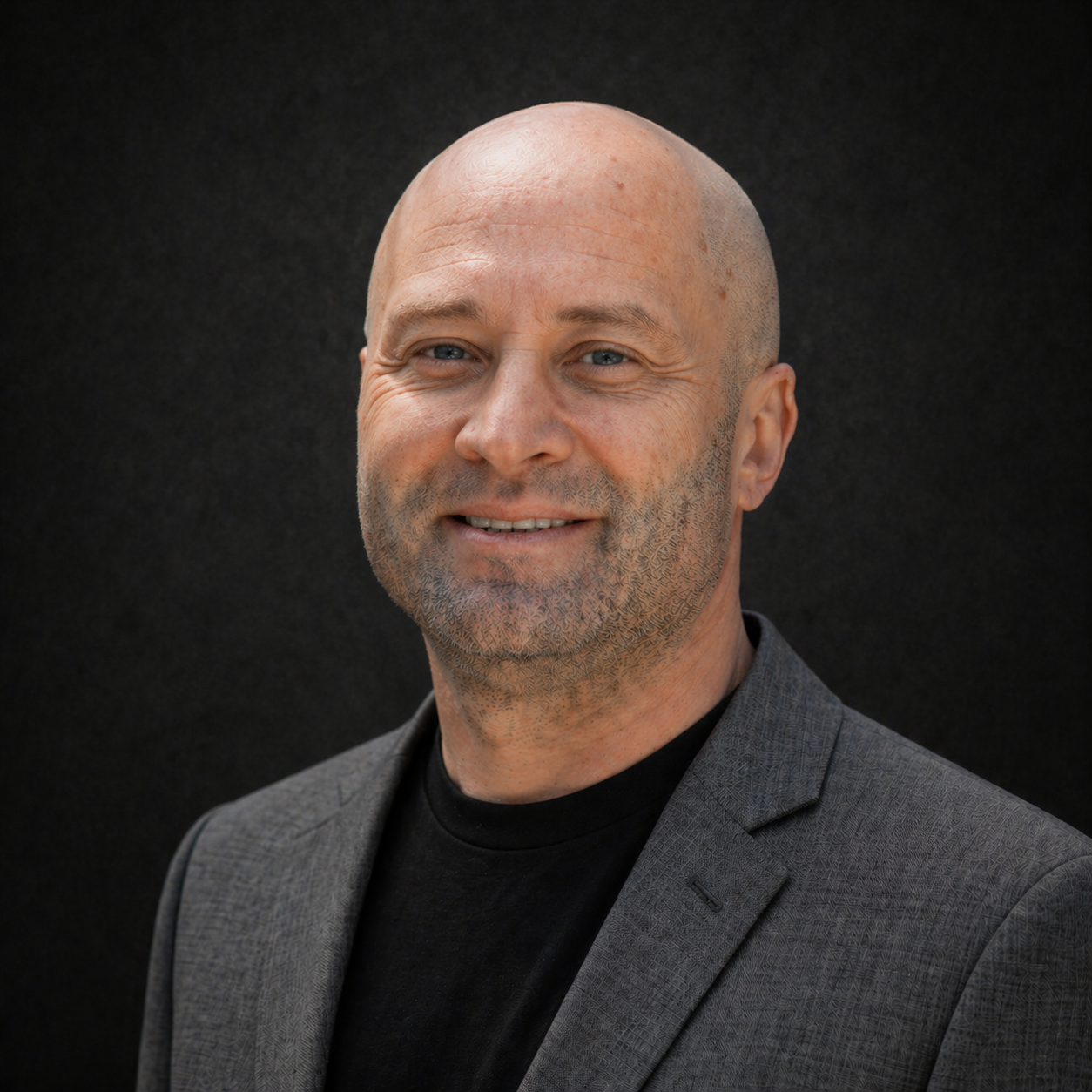 A bald man outdoors smiling at the camera with a house and trees in the background.