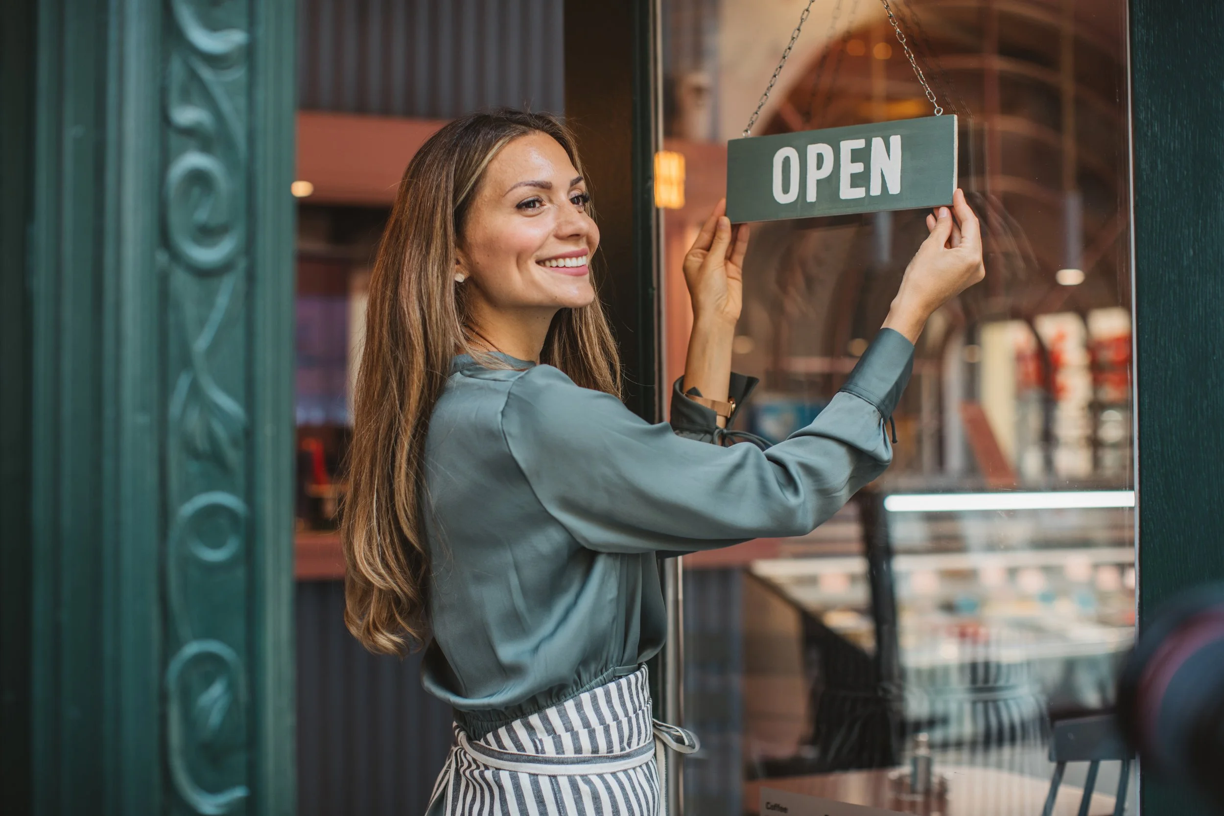A woman with long brown hair smiling and hanging a green 'OPEN' sign on a door.