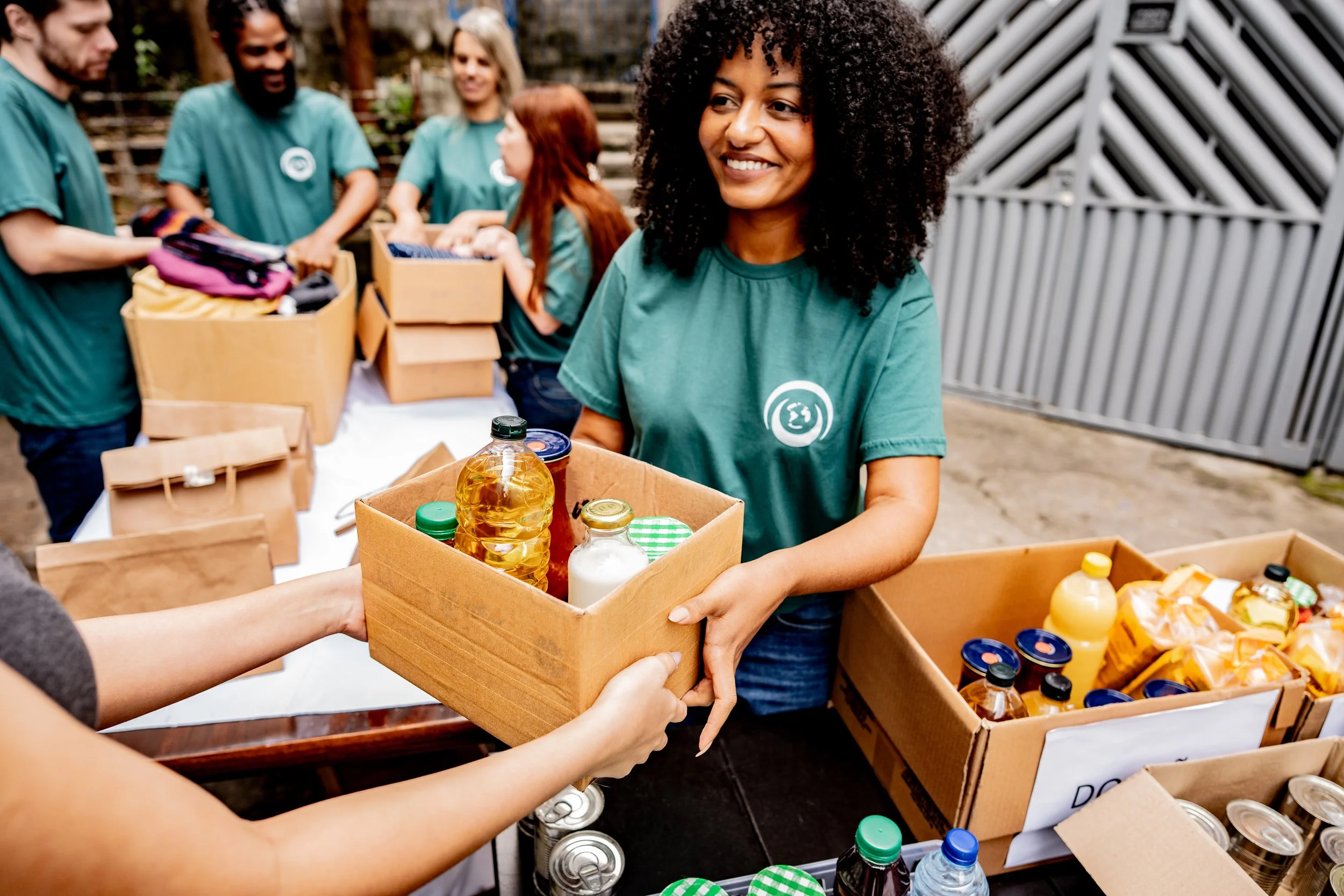 A woman with curly hair and a teal T-shirt is smiling while receiving a box of food items from volunteers. The volunteers are in teal T-shirts and are seen in the background handing out boxes with various food supplies, including bottled drinks and canned goods. The setting appears to be outdoors, possibly at a community food drive or distribution event.