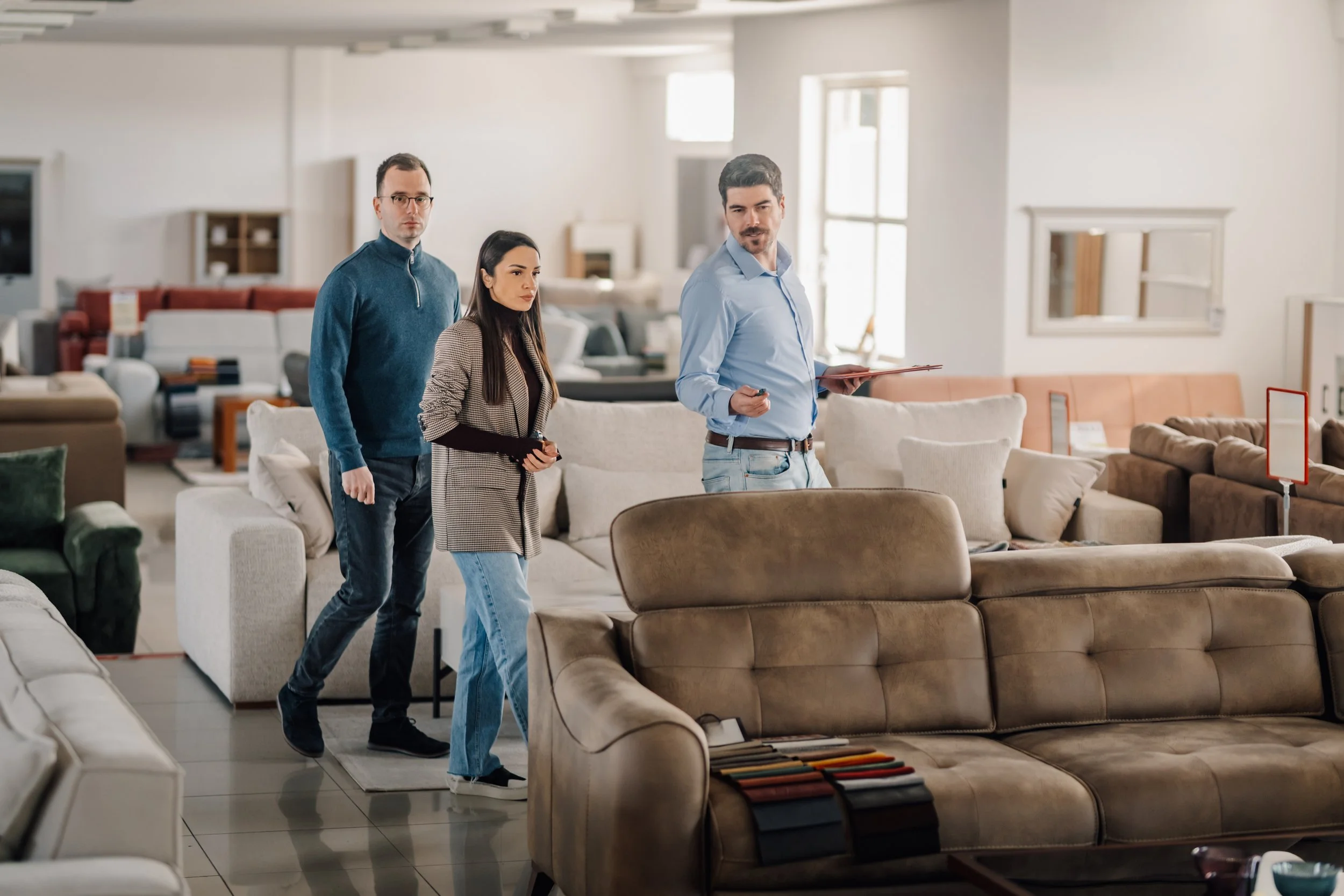 Three people are shopping for sofas in a furniture store. They are walking among display couches in a well-lit showroom.