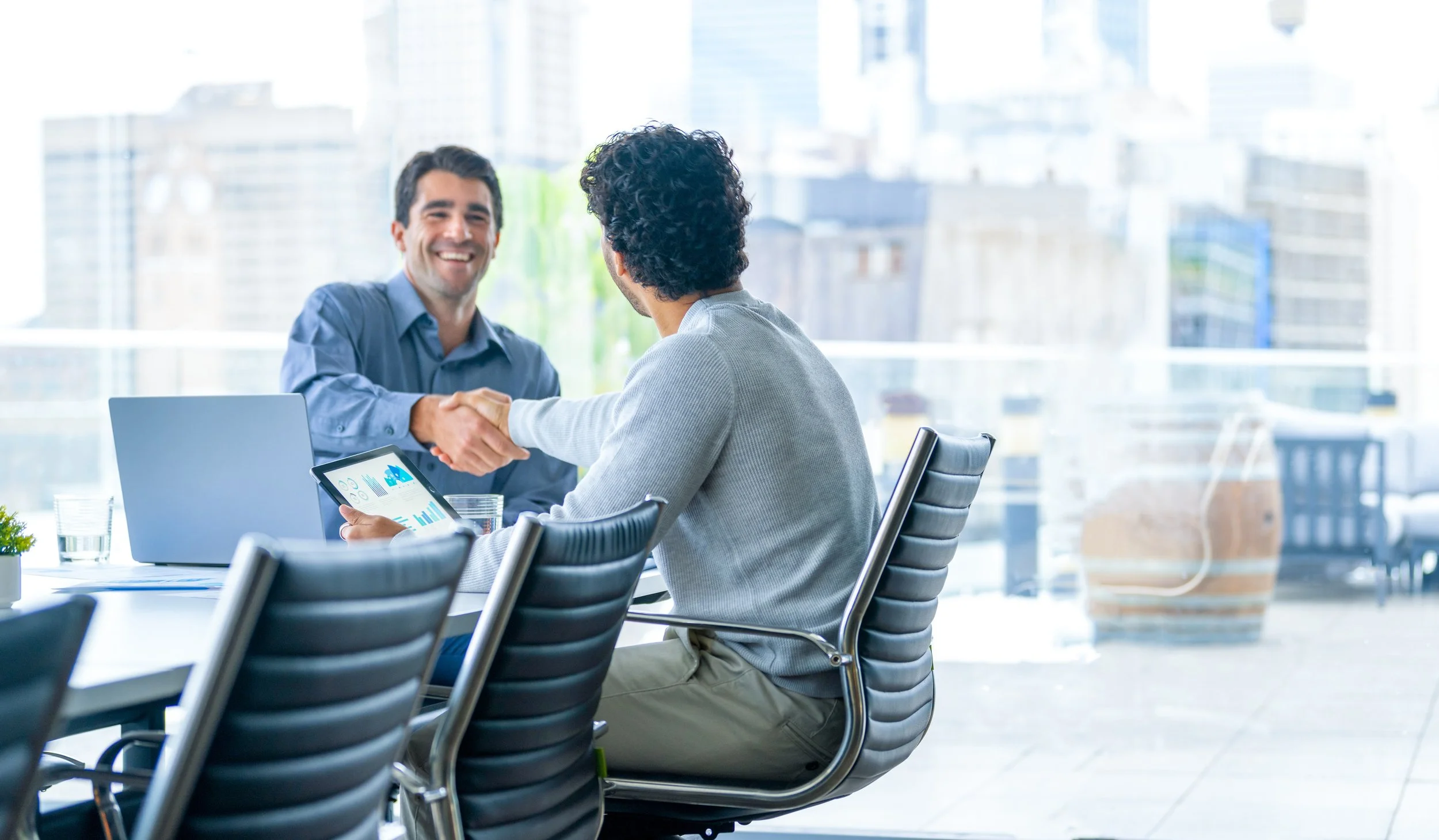 Two men in business attire shake hands across a conference table in a modern office with cityscape views.