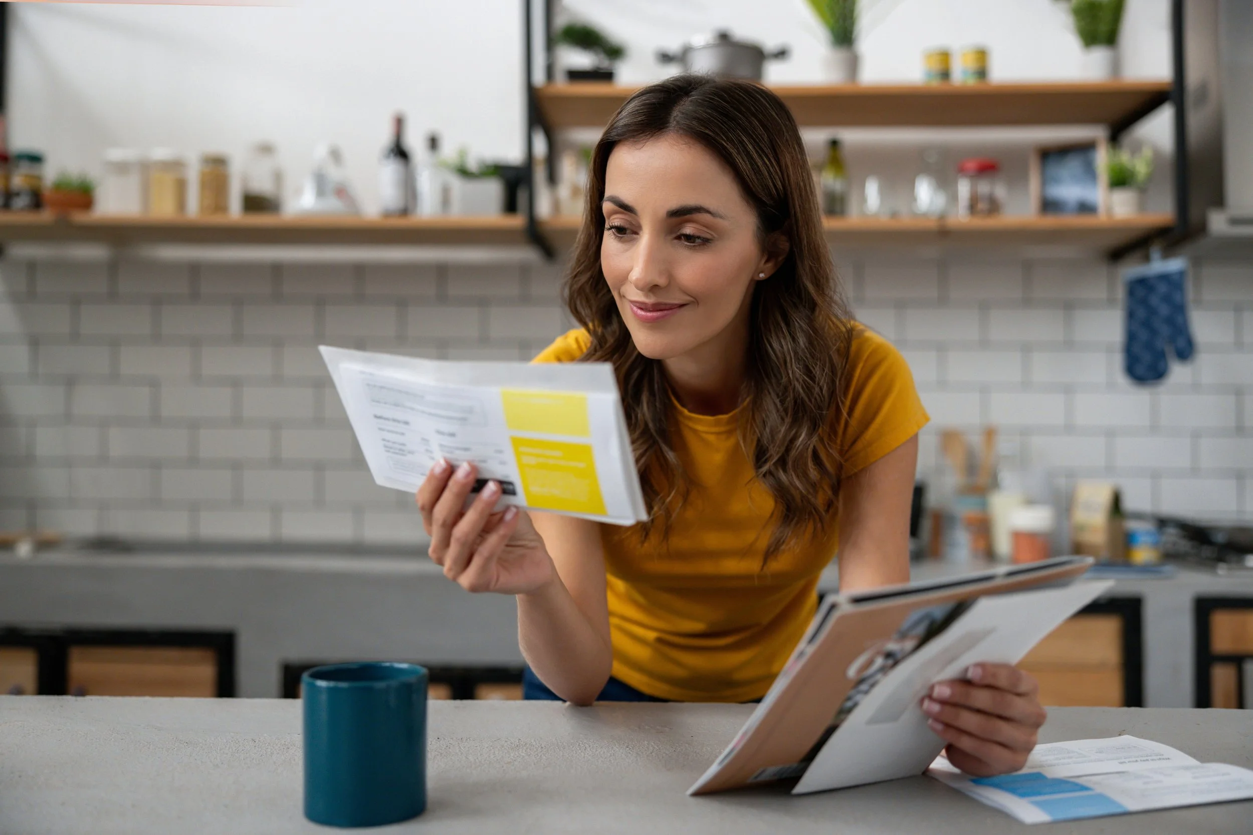 A woman with brown hair wearing a yellow shirt looks at a yellow and gray booklet while sitting at a kitchen counter with a blue mug nearby. The background features open shelves with jars, bottles, and plants.
