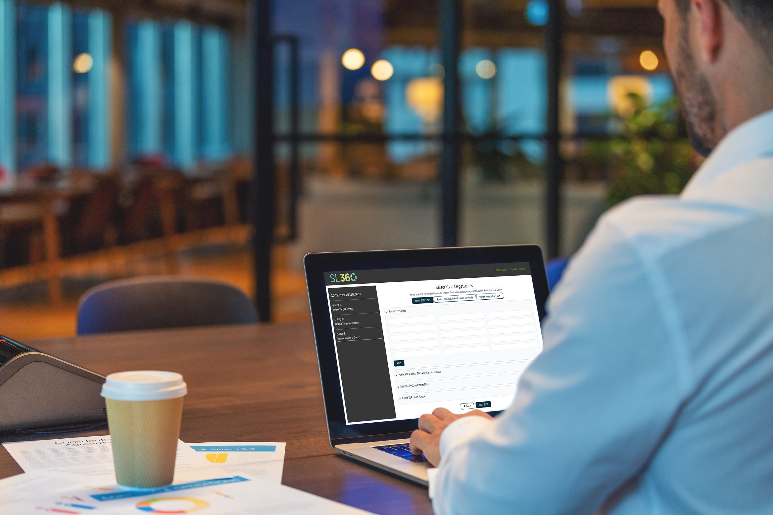 A man in a white shirt sitting at a wooden table using a laptop with a digital business application on the screen. There are papers and a disposable coffee cup on the table, and blurred background of a modern cafe or workspace with warm lighting.