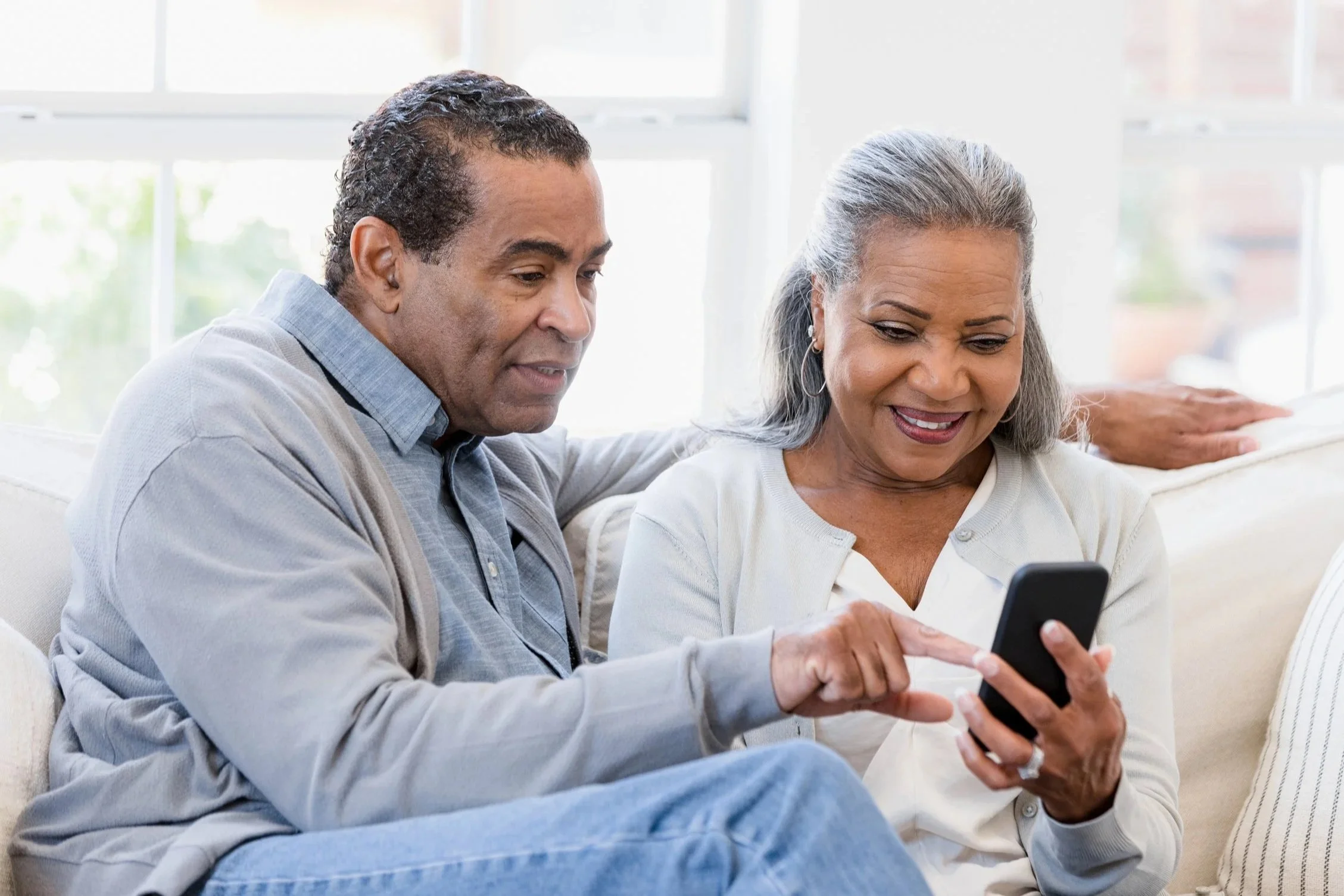 A middle-aged man and woman sitting on a sofa, looking at a smartphone together in a bright, airy room with large windows.