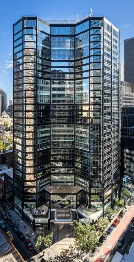 Tall modern glass office building in a city, reflecting surrounding skyscrapers and blue sky.