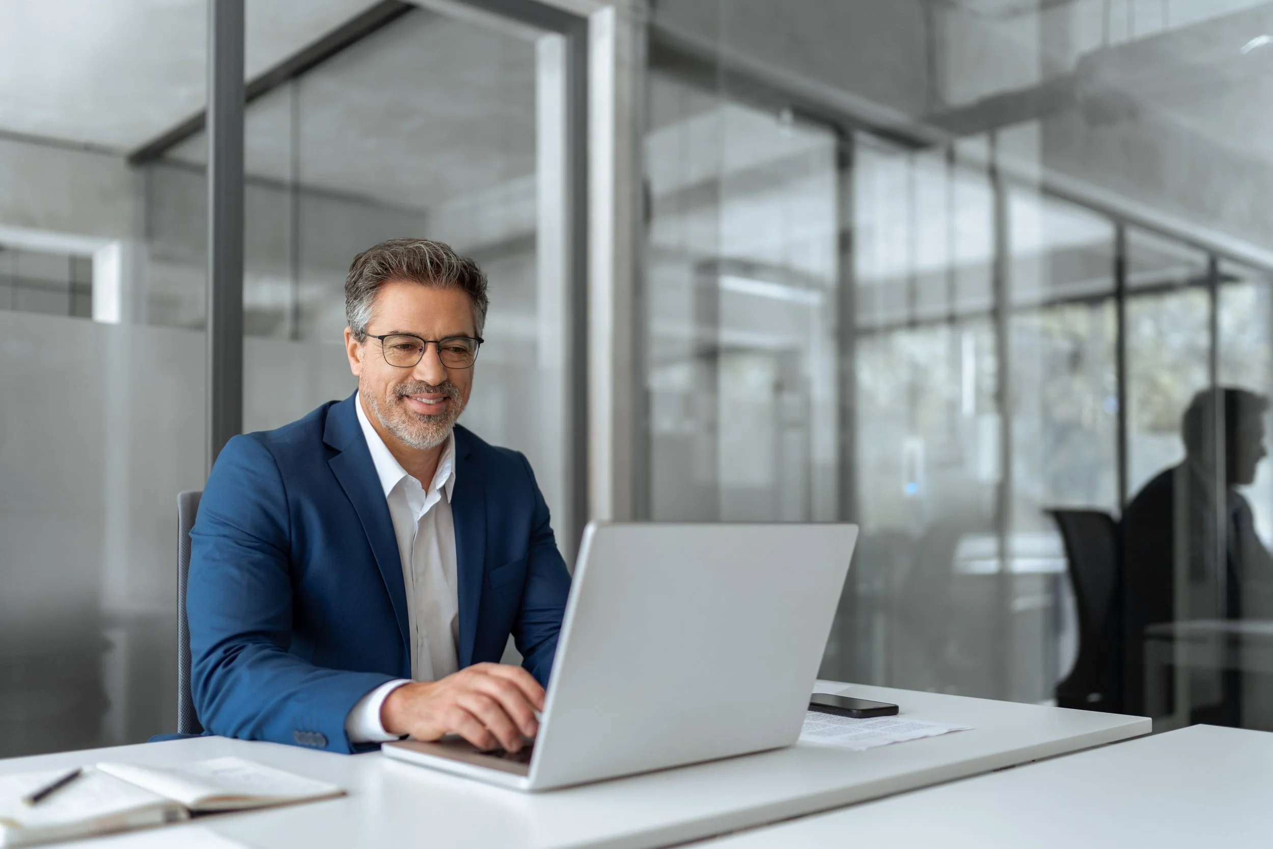 A middle-aged man with gray hair, glasses, and a beard, wearing a blue suit and white shirt, is sitting at a desk working on a laptop in a modern office environment with glass walls and other people working in the background.