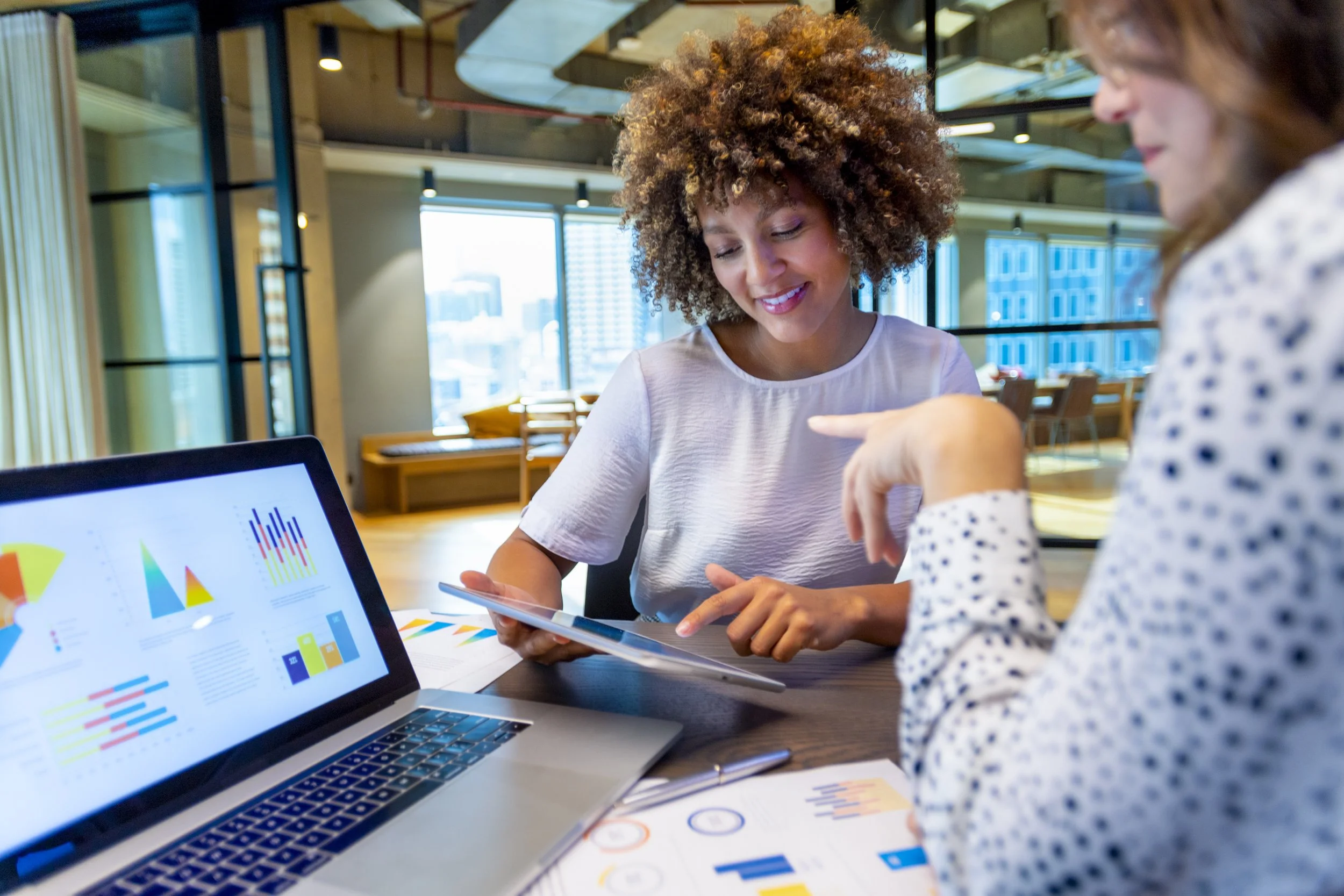 Two women at a business meeting or discussion, one holding a tablet and the other pointing at a chart on a laptop screen with colorful graphs and data.