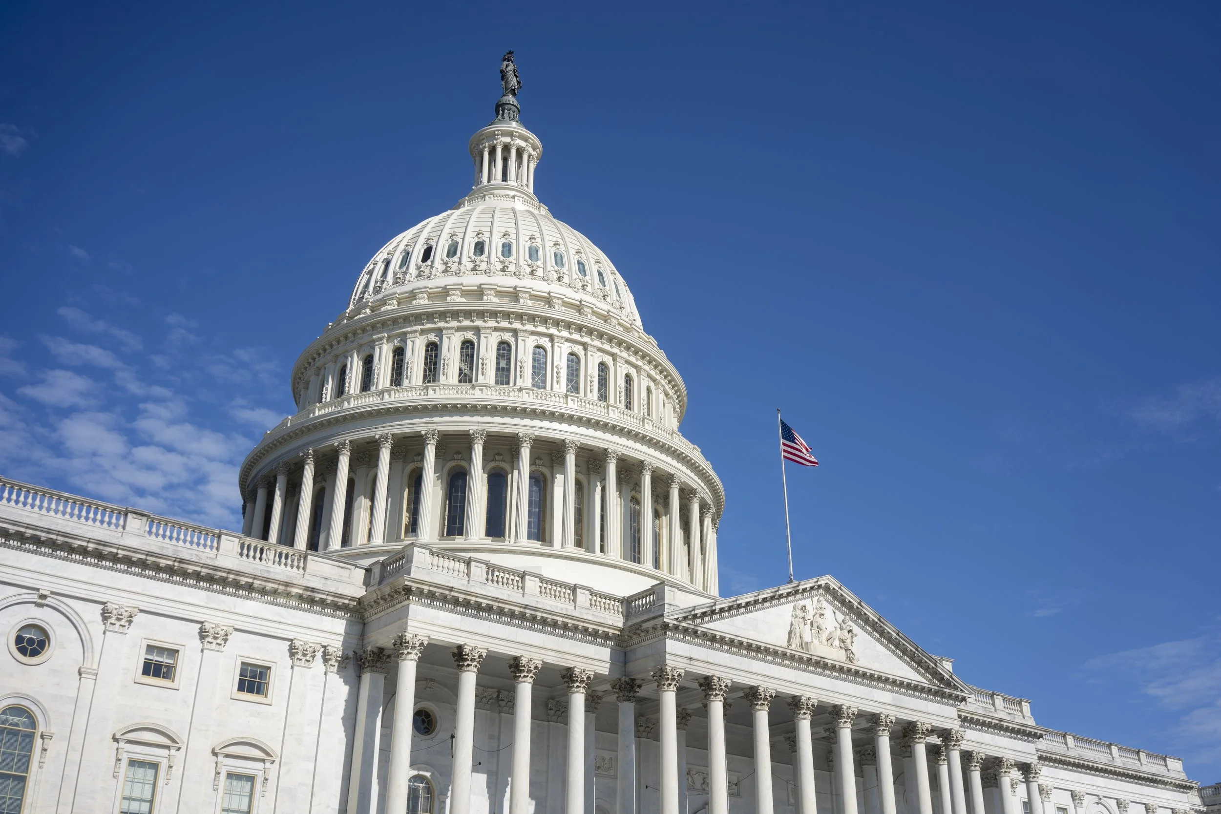 The United States Capitol building under a bright blue sky, with an American flag flying in front.