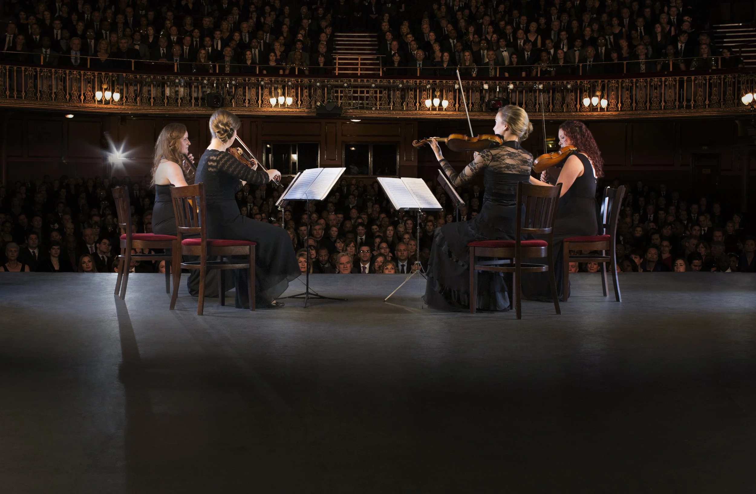 A string quartet performing on stage in front of an audience in a concert hall with ornate railings and multiple levels of spectators.