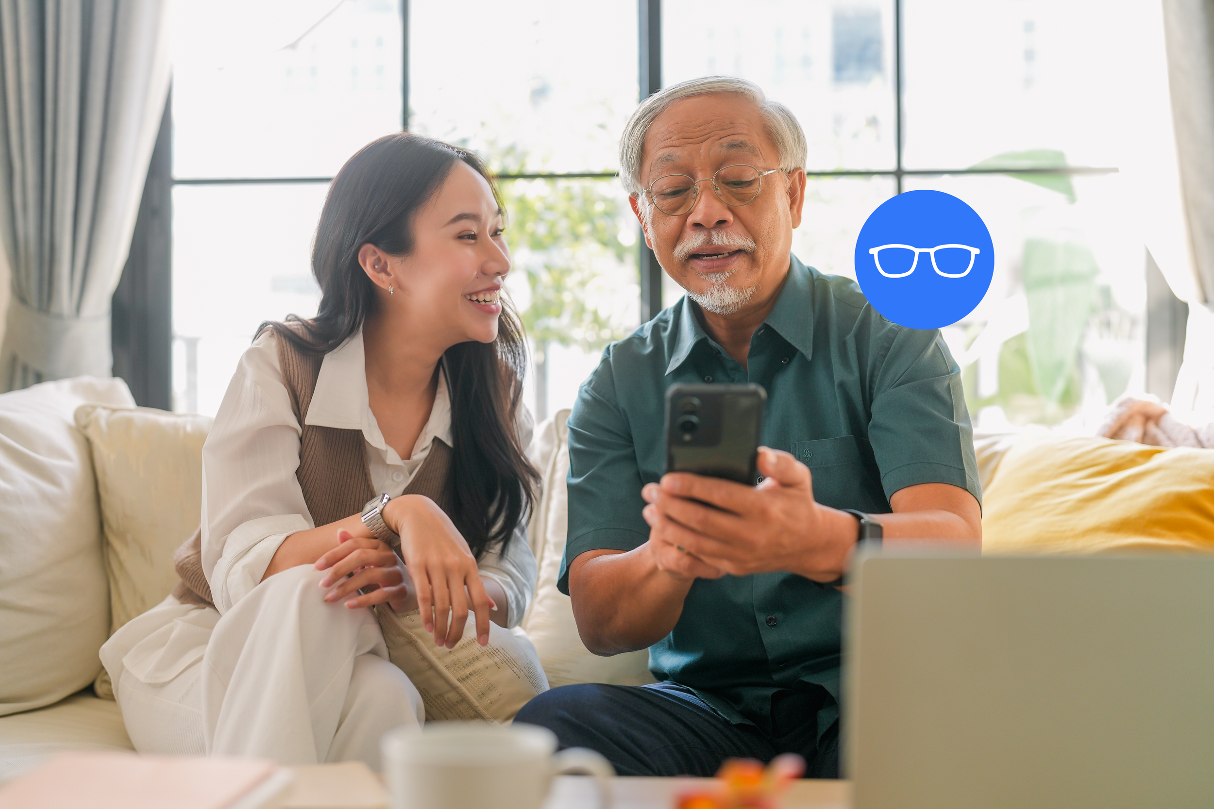 A young woman and an older man are sitting together on a sofa, looking at a smartphone. The young woman is smiling and the older man is wearing glasses, appears to be explaining something, and both are engaged with the phone. There is a large window behind them with sunlight and greenery outside.