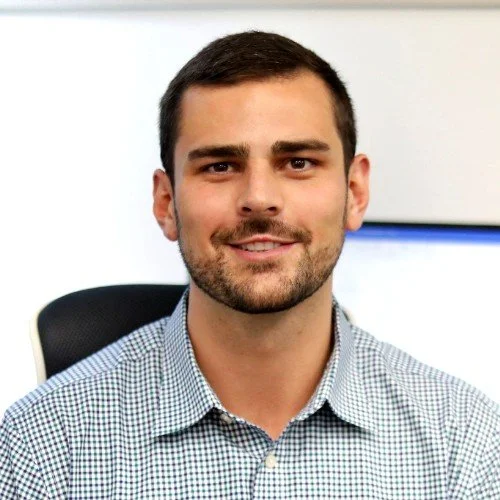 Portrait of a young man with short dark hair and a beard, smiling, wearing a checkered shirt, seated on an office chair with a blue and white background.
