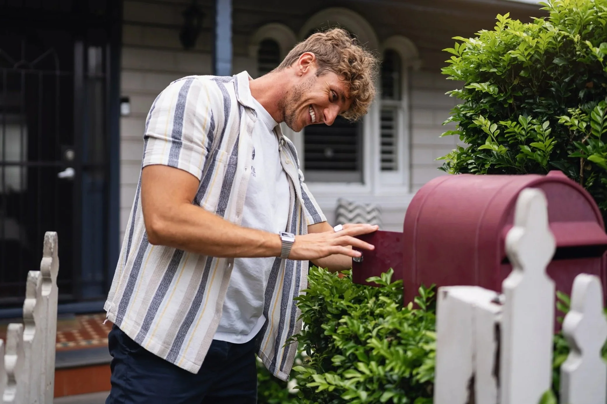 A man with curly hair and a beard smiling as he looks down at a mailbox in front of a house with beige siding and a white picket fence, surrounded by green bushes.