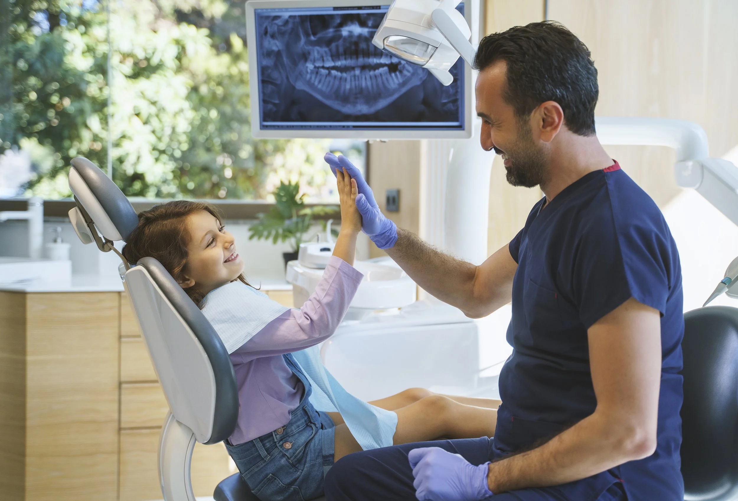 A young girl sitting in a dental chair, smiling and giving a high-five to a smiling male dentist or dental hygienist dressed in blue scrubs and wearing gloves, with a dental X-ray displayed on a monitor in the background.