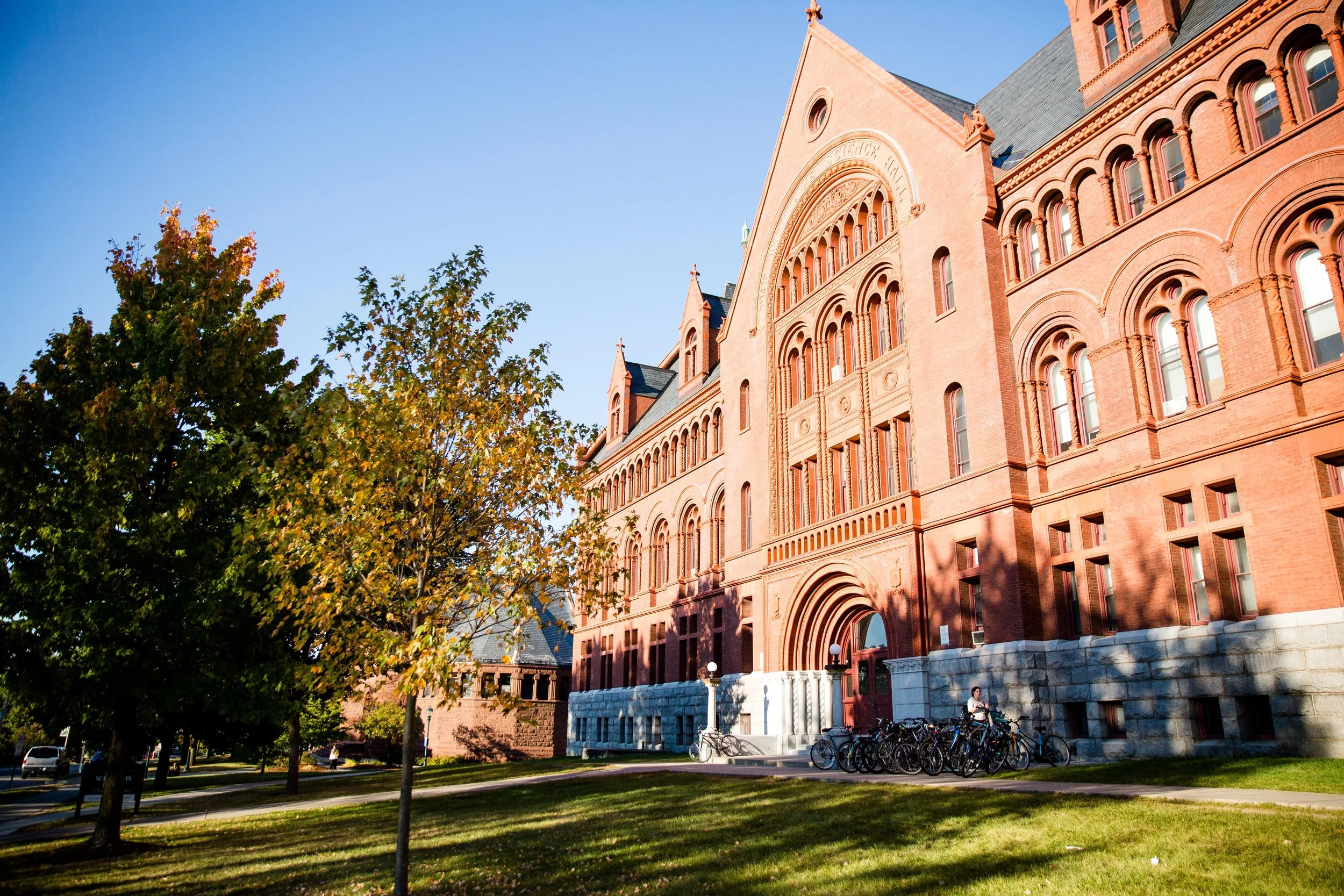 A large historic brick building with arched windows and a  steep roof, surrounded by trees with green and yellow leaves, under a clear blue sky.