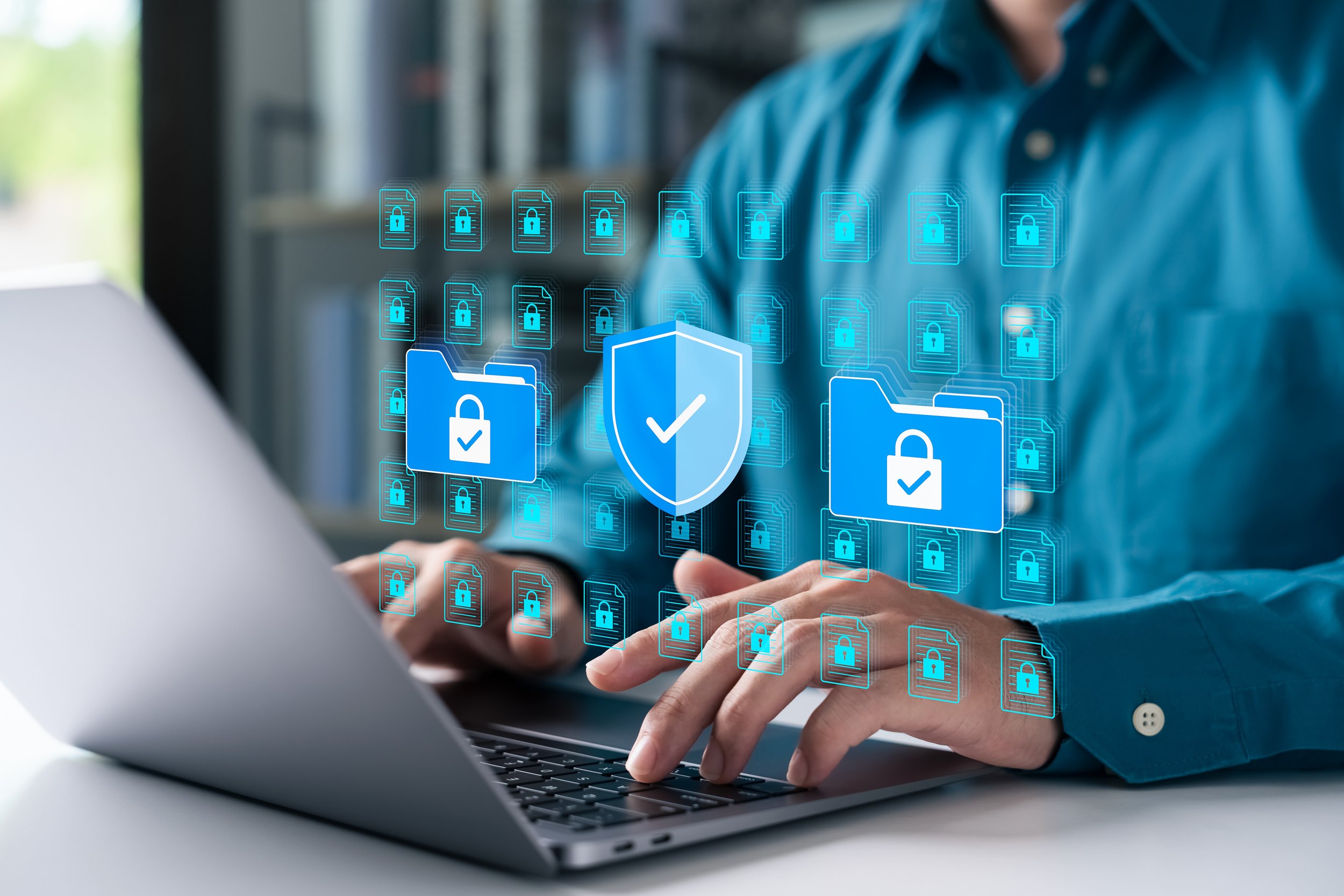 Person in blue shirt using a laptop with digital icons of locked files and security shields related to cybersecurity.