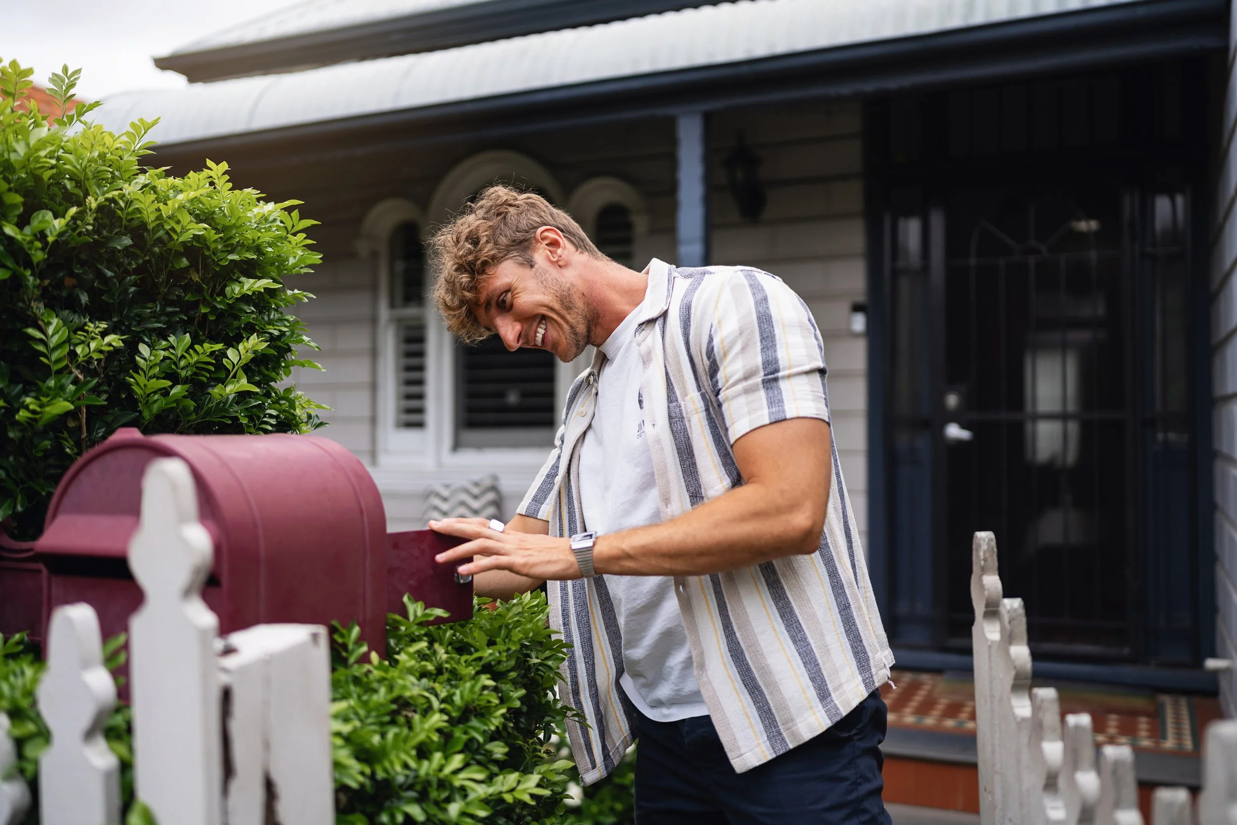 A man smiling while opening a red mailbox on his porch, with greenery and white picket fence in the foreground.