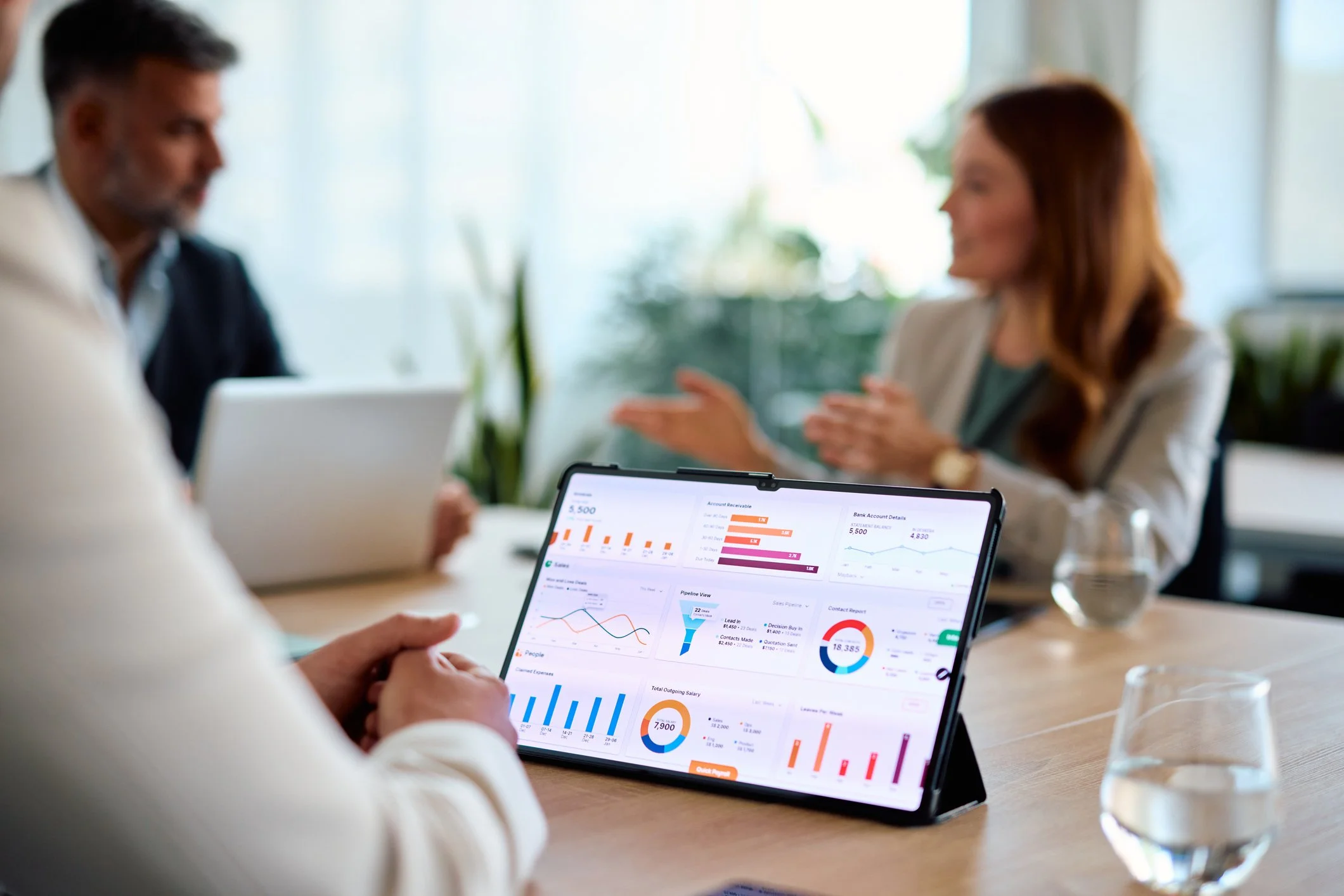 People in a business meeting discussing financial charts on a tablet and laptop in a bright office environment.
