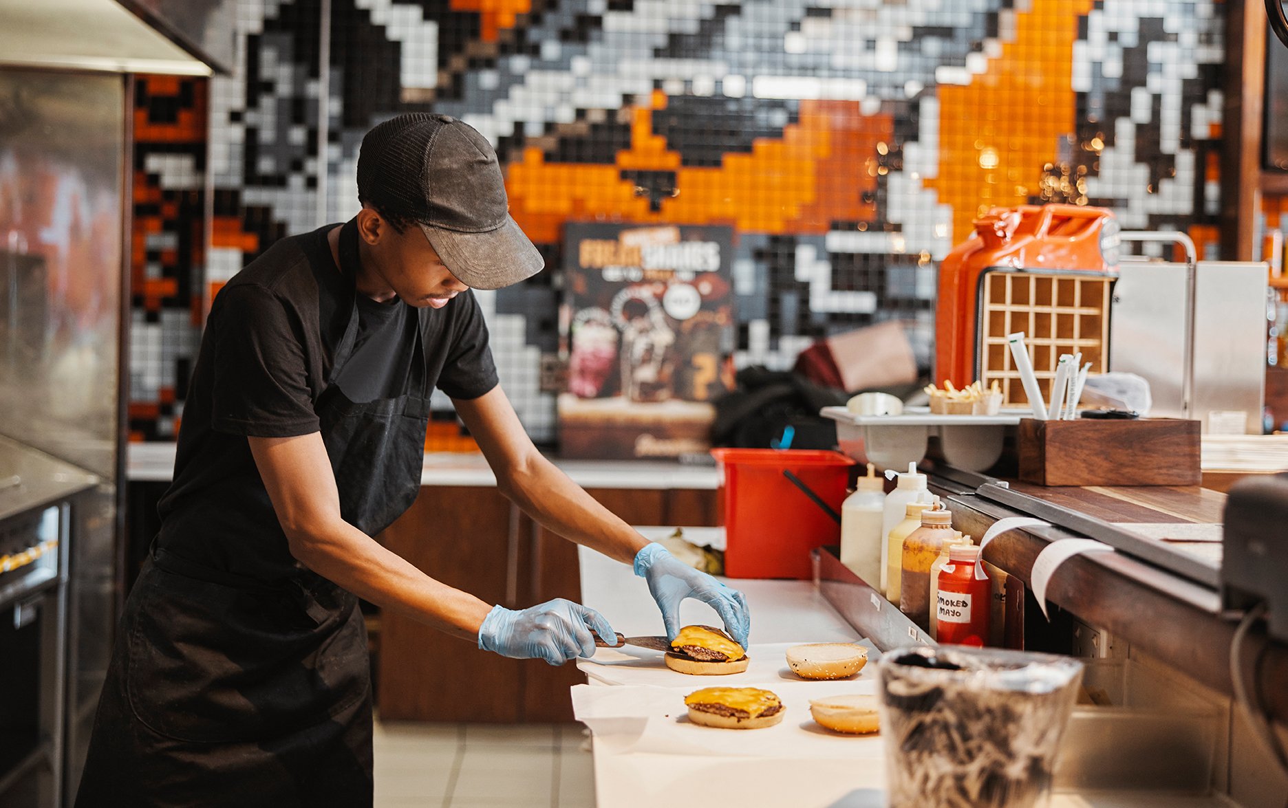A fast food worker wearing a cap, black shirt, black apron, and gloves prepares burgers in a restaurant kitchen with modern black, white, and orange tiled walls.
